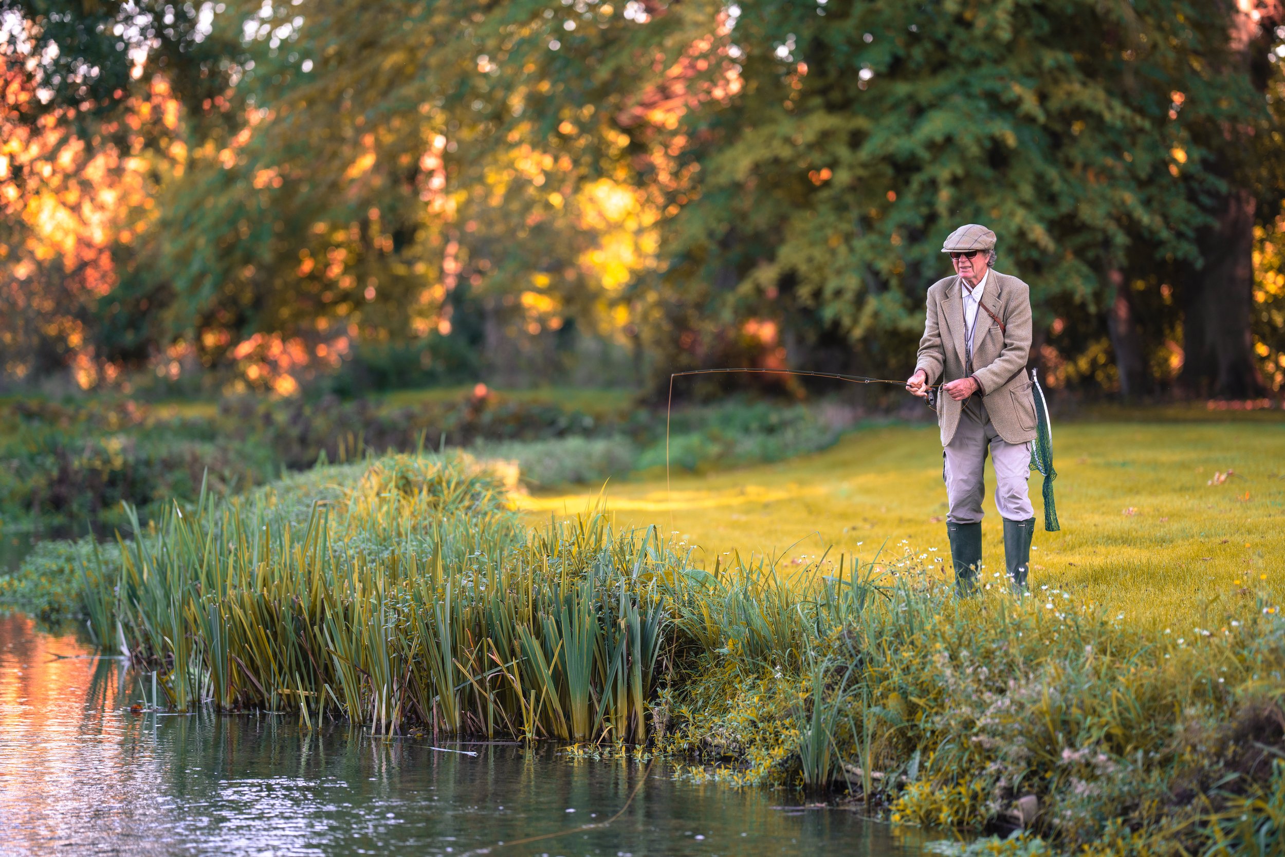 An elderly man fishing in a pond surrounded by trees during autumn with colorful foliage.