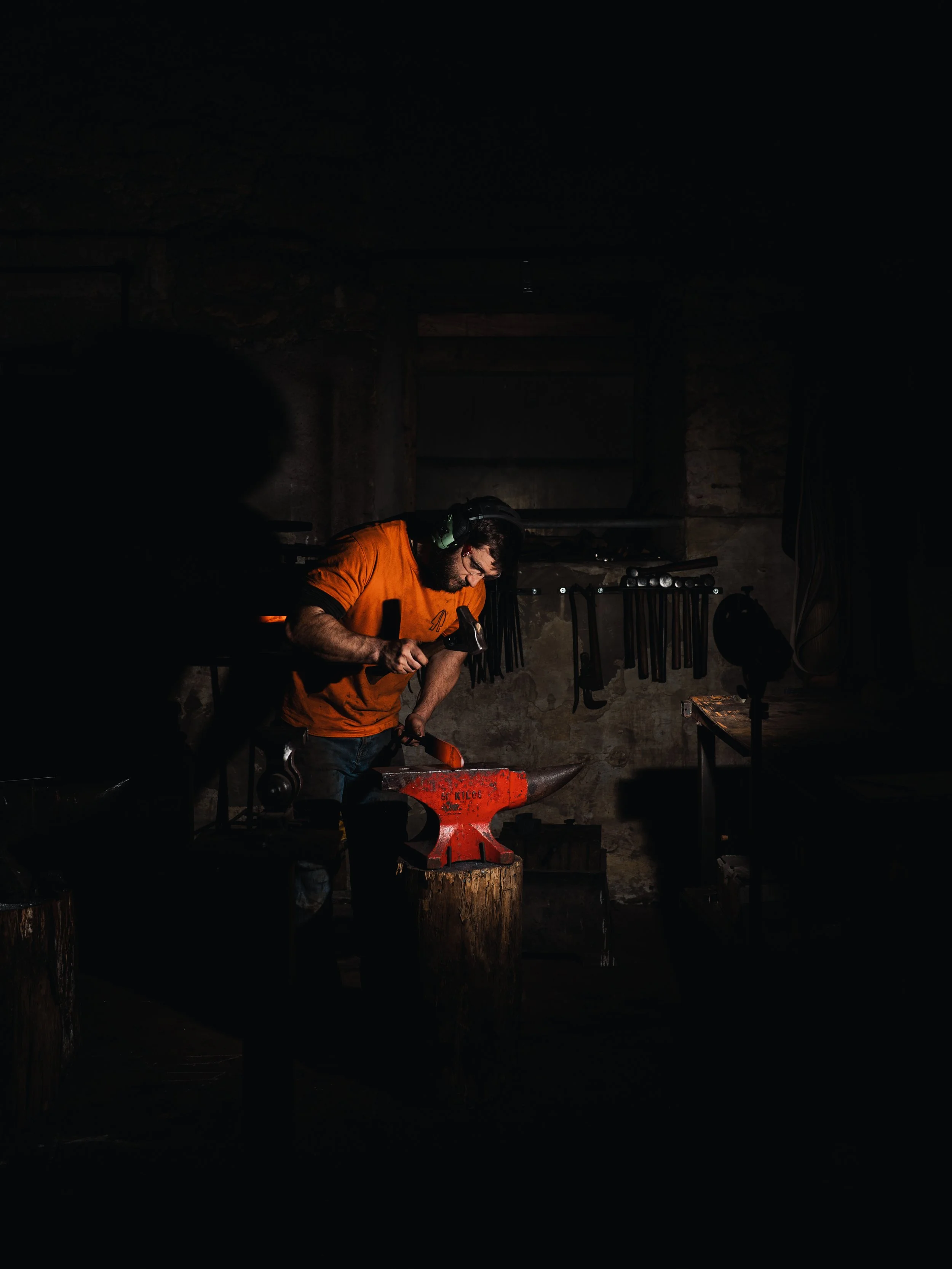 Blacksmith working with a hammer on an anvil in a dark workshop, wearing safety gear.