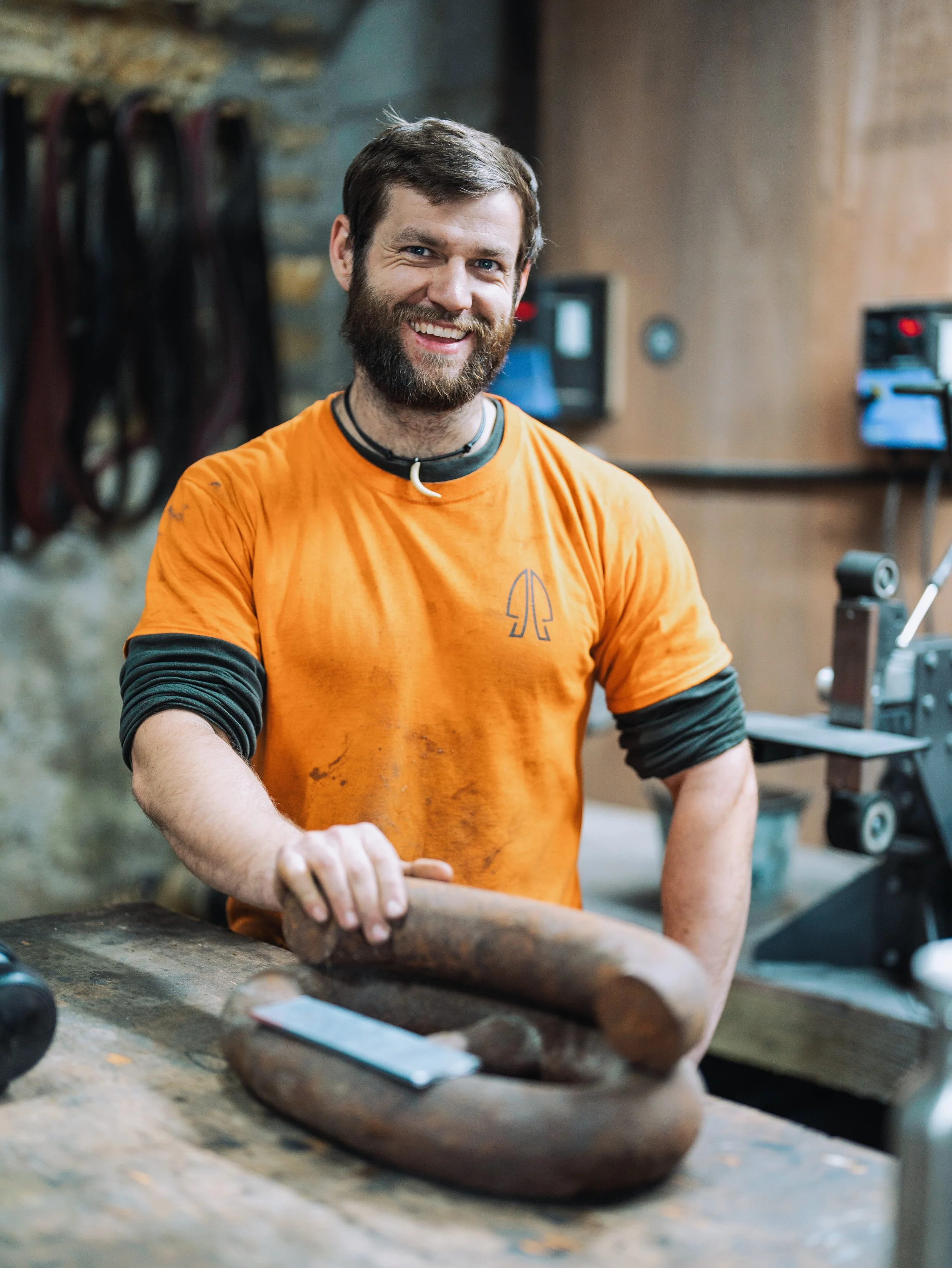 A man with a beard and mustache wearing an orange t-shirt is crafting a piece of wood in a woodworking shop. The background shows woodworking tools hanging on the wall and equipment used for woodworking.