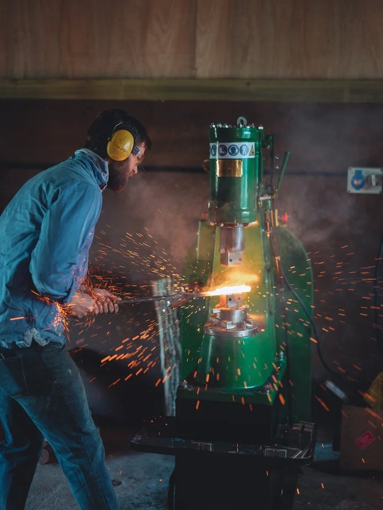 A man wearing ear protection and glasses is welding metal in a workshop with sparks flying.