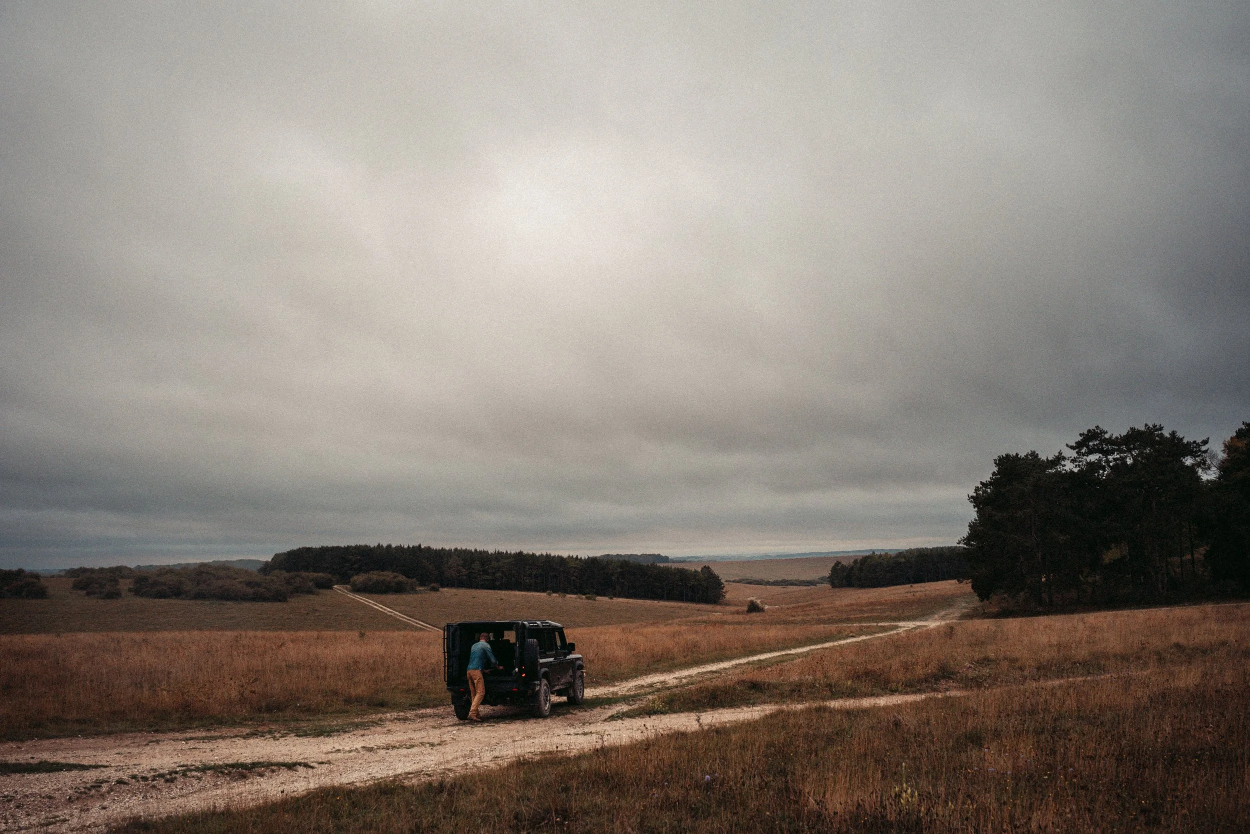 A person loading items into the back of a black pickup truck on a rural dirt road, with an expansive field and trees under a cloudy sky.