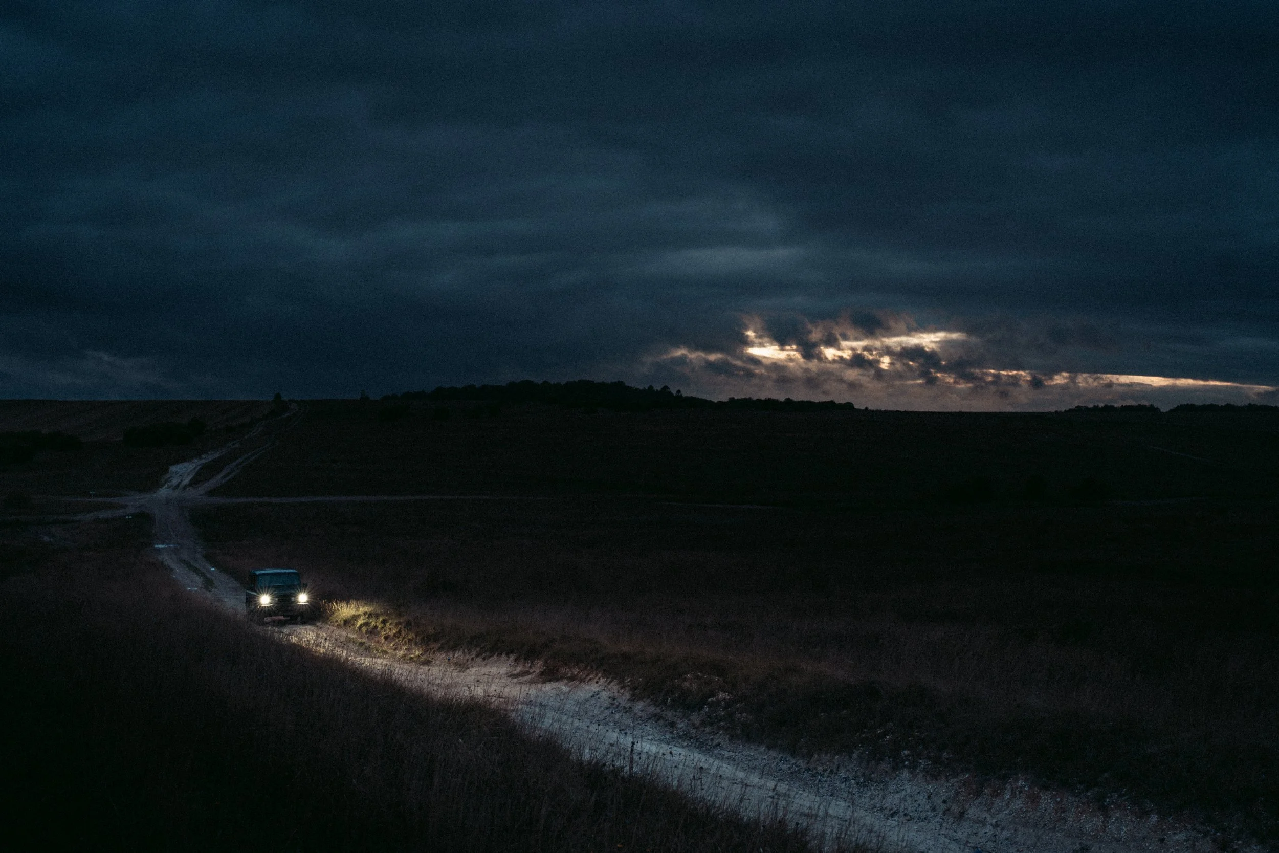 Dark, cloudy sky over a landscape with a dirt road and rolling hills. A car with headlights on is on the road, heading toward the viewer.