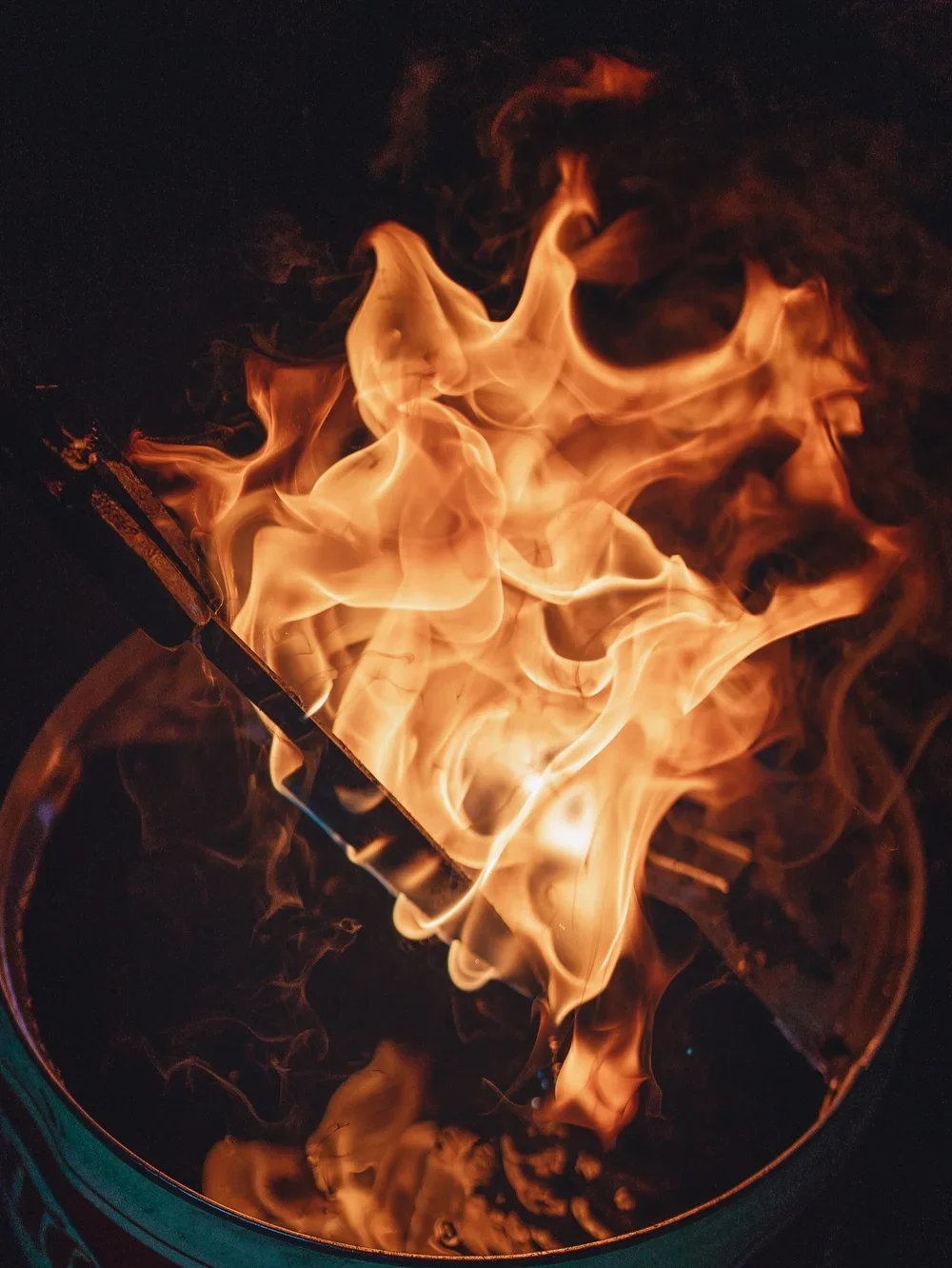Flames burning inside a metal container, with a metal tool or stick partially visible amidst the fire.