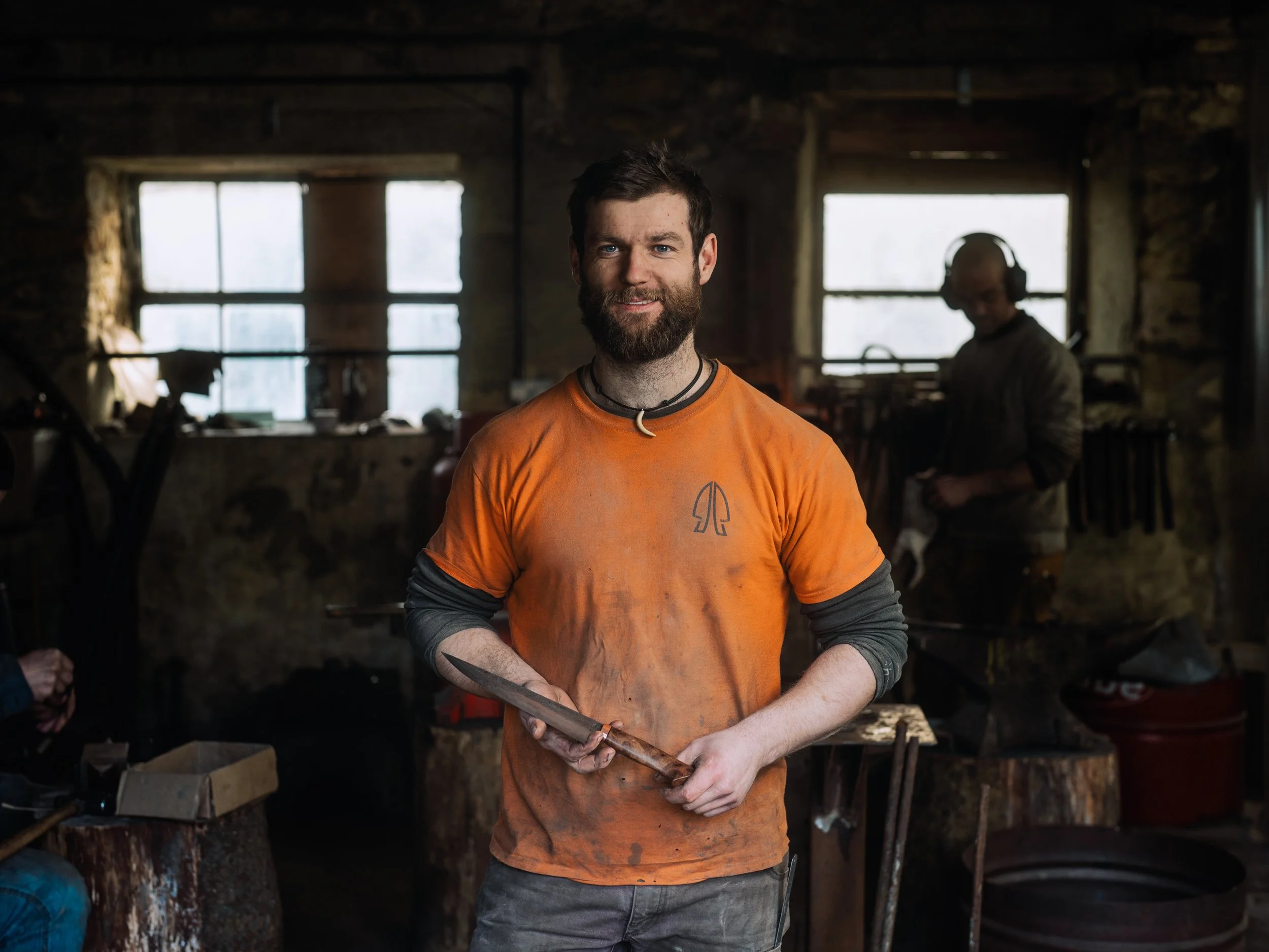 A man with a beard and dark hair wearing an orange shirt with a small logo at the chest, holding a metal tool, standing in a workshop with wooden and stone walls, and another person in the background wearing headphones, working at a bench.