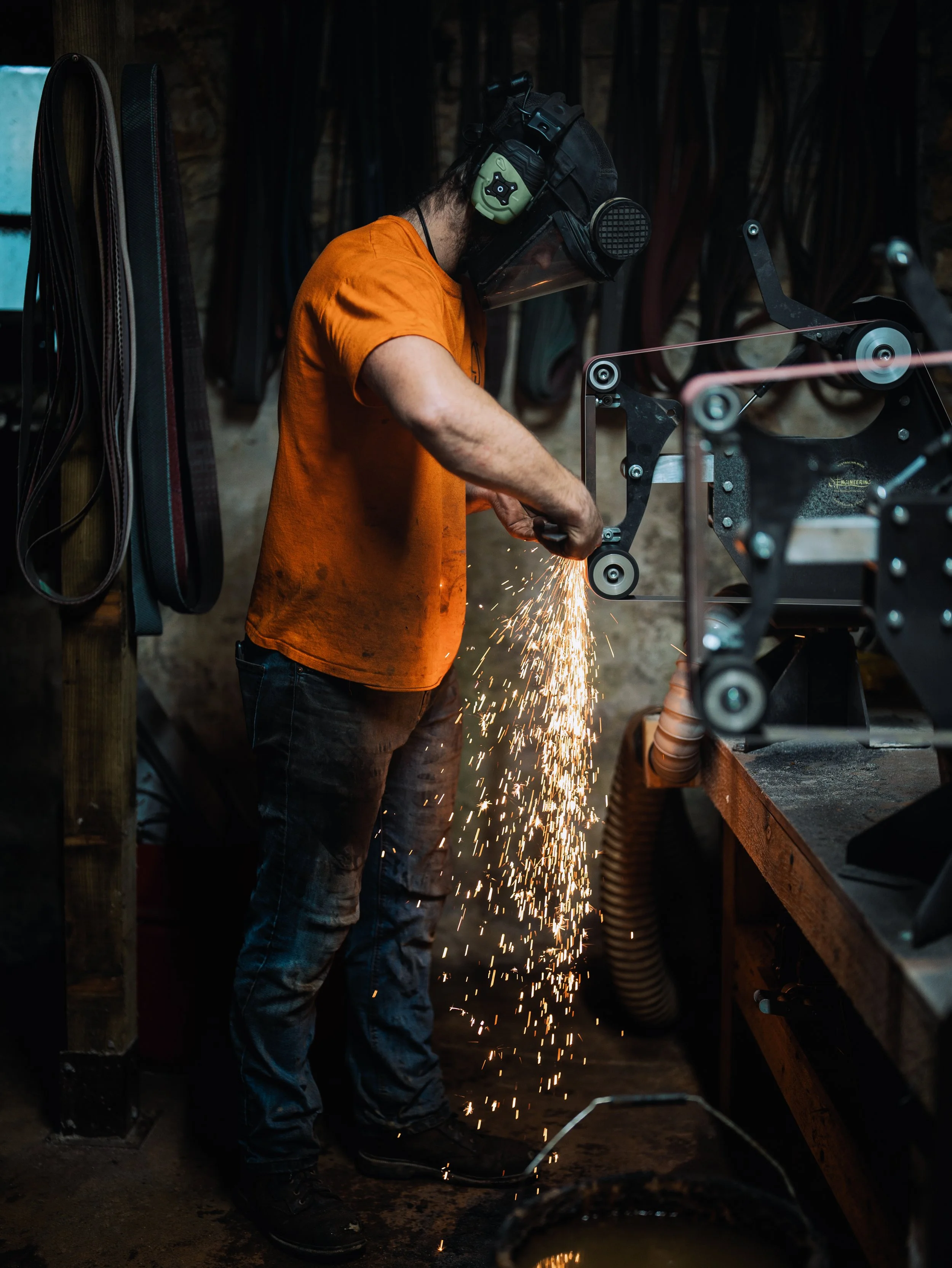 A person wearing safety gear, including a face shield, welding helmet, green earmuffs, and an orange shirt, is welding metal with sparks flying in a workshop with various tools and materials in the background.