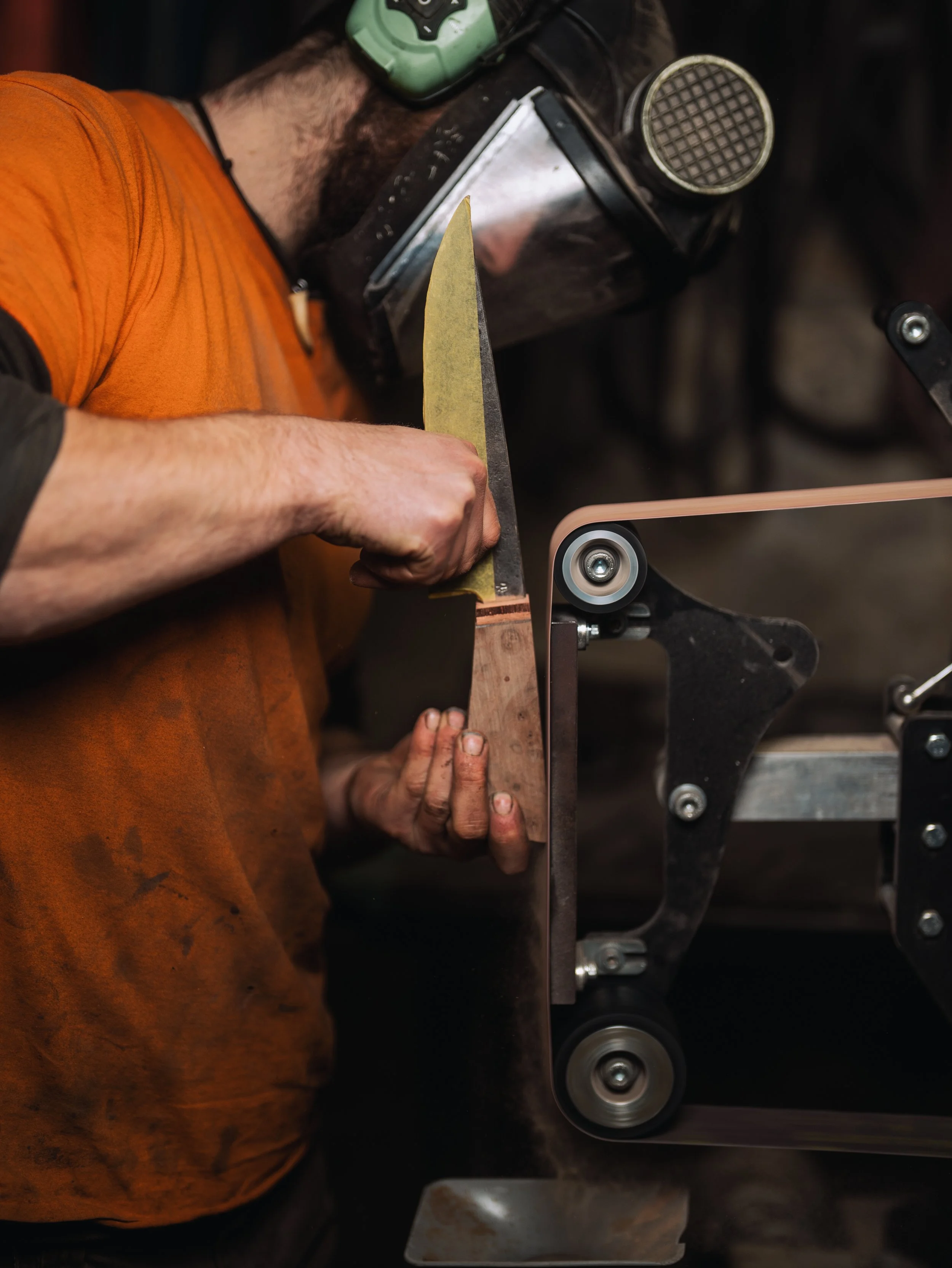 A person wearing an orange shirt and safety goggles operates a belt sander to shape or smooth a piece of wood, with a focus on the woodworking process.