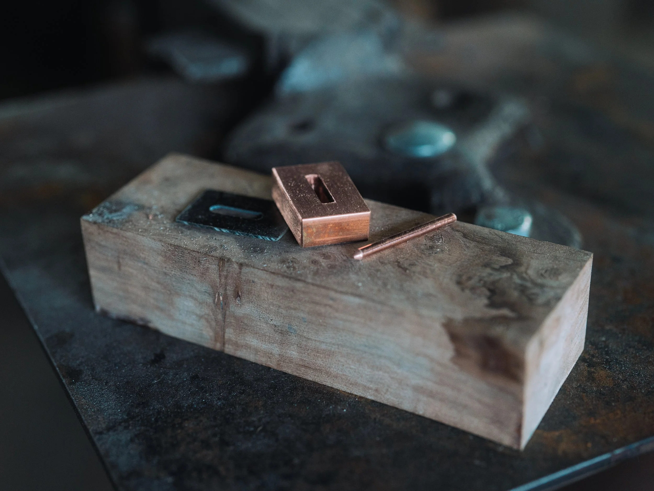 A small copper-colored metal component on a workbench with a black and gray background, possibly in a workshop or metalworking environment.