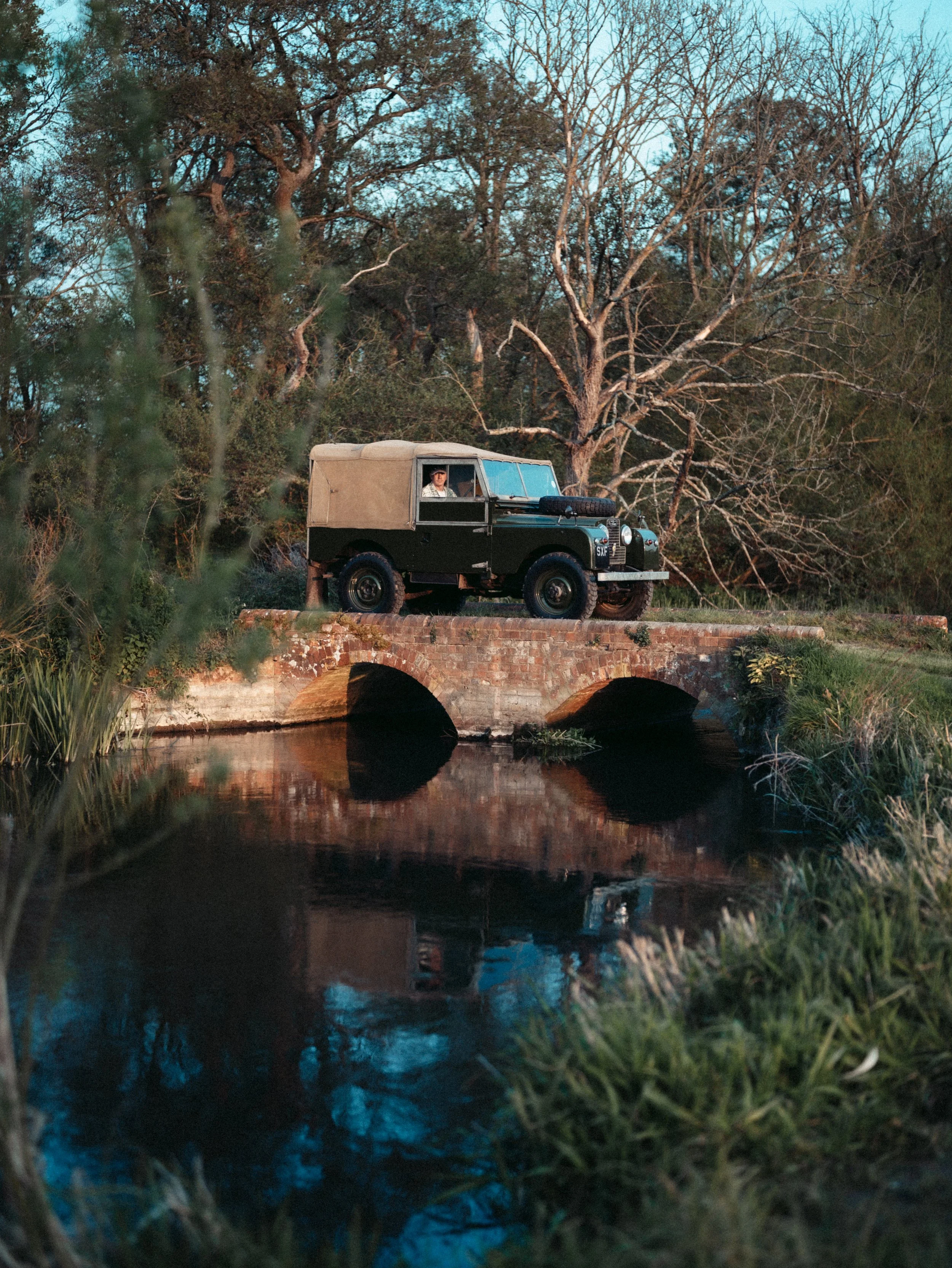 A vintage off-road vehicle with a beige canopy and a spare tire on the hood, parked on a small brick bridge over a river, amidst trees with barren branches and some greenery, during dusk or early evening.