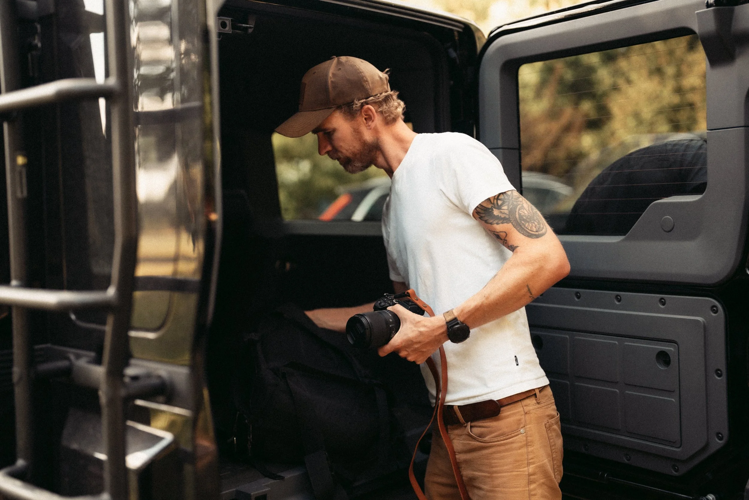 A man with tattoos on his arm wearing a brown cap and a white t-shirt is looking inside an open black vehicle, possibly a Jeep, with trees visible in the background.