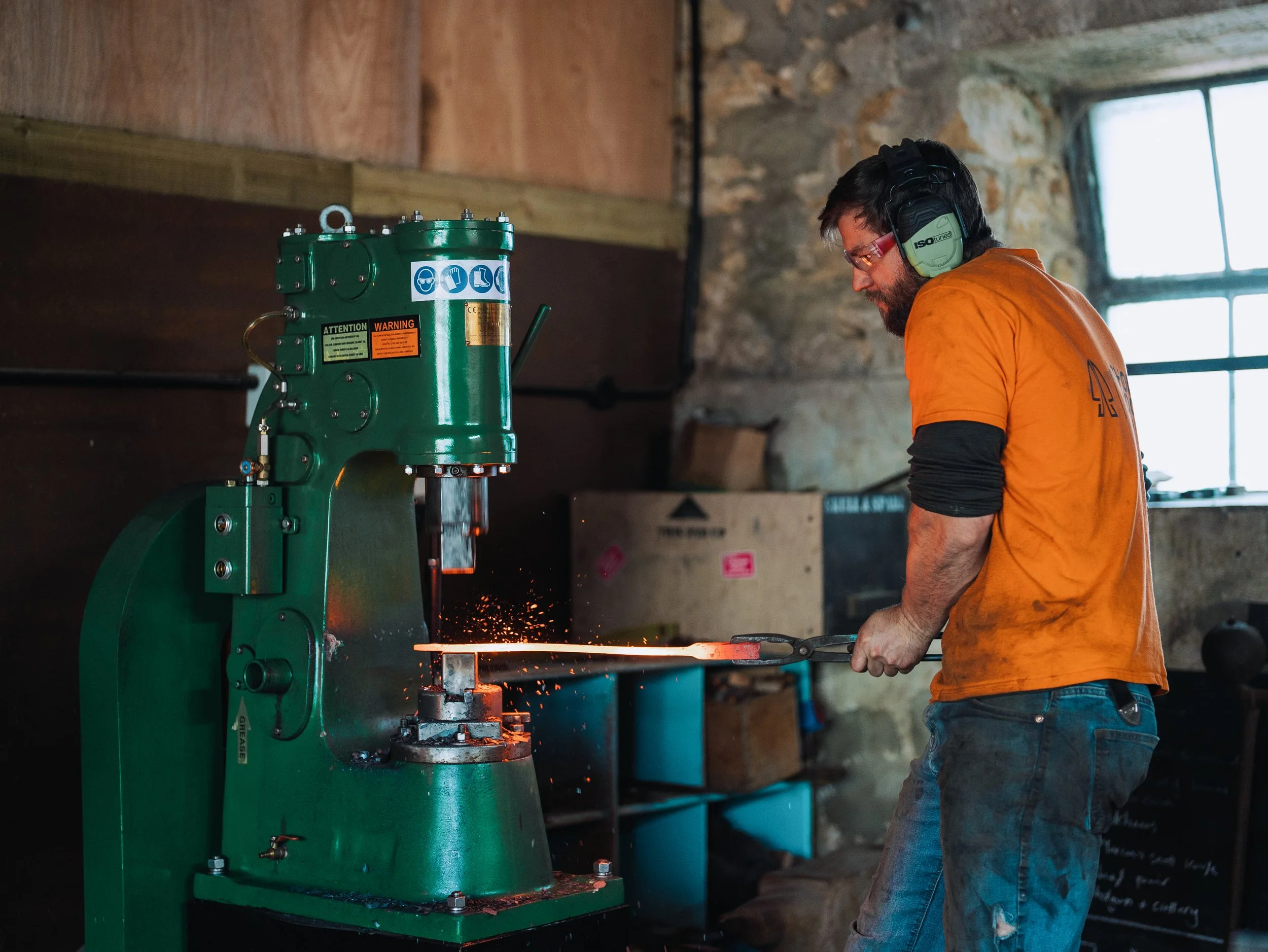 A man wearing safety glasses, earmuffs, an orange t-shirt, and jeans is working with a green industrial machine in a workshop with a stone wall and window in the background.