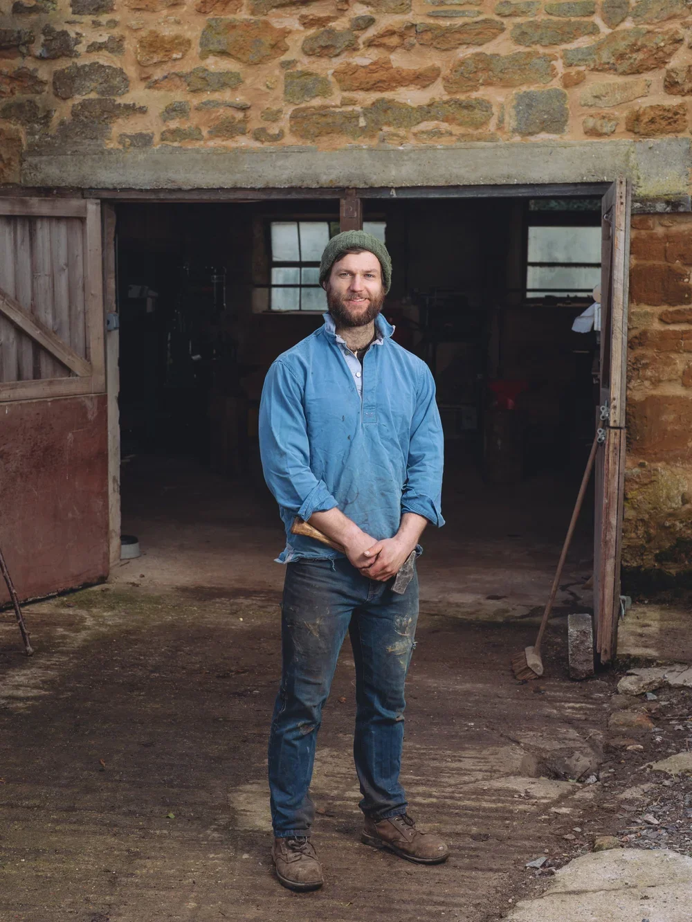 A man standing in front of a rustic barn with open sliding doors, wearing a green beanie and a blue work shirt, smiling at the camera.