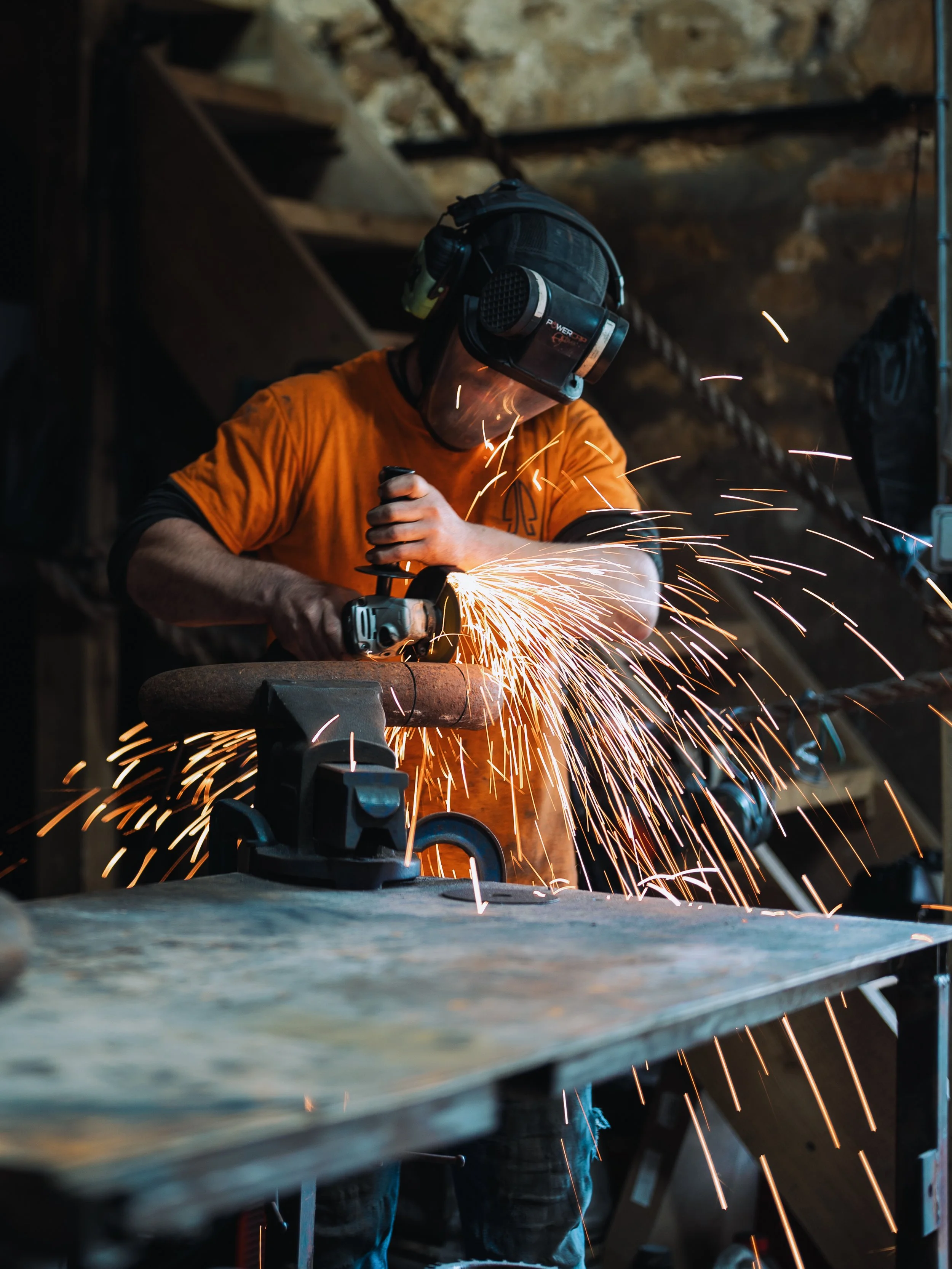 A person welding metal with sparks flying, wearing safety gear including a helmet, face shield, and protective gloves in an industrial workshop.