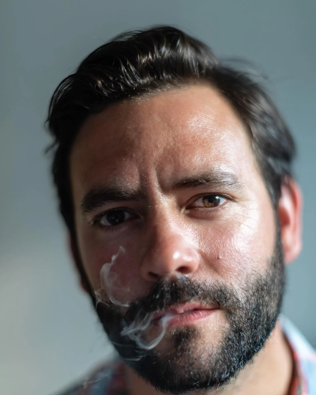 Close-up of a man with dark hair and a beard, looking directly at the camera with smoke near his lips.
