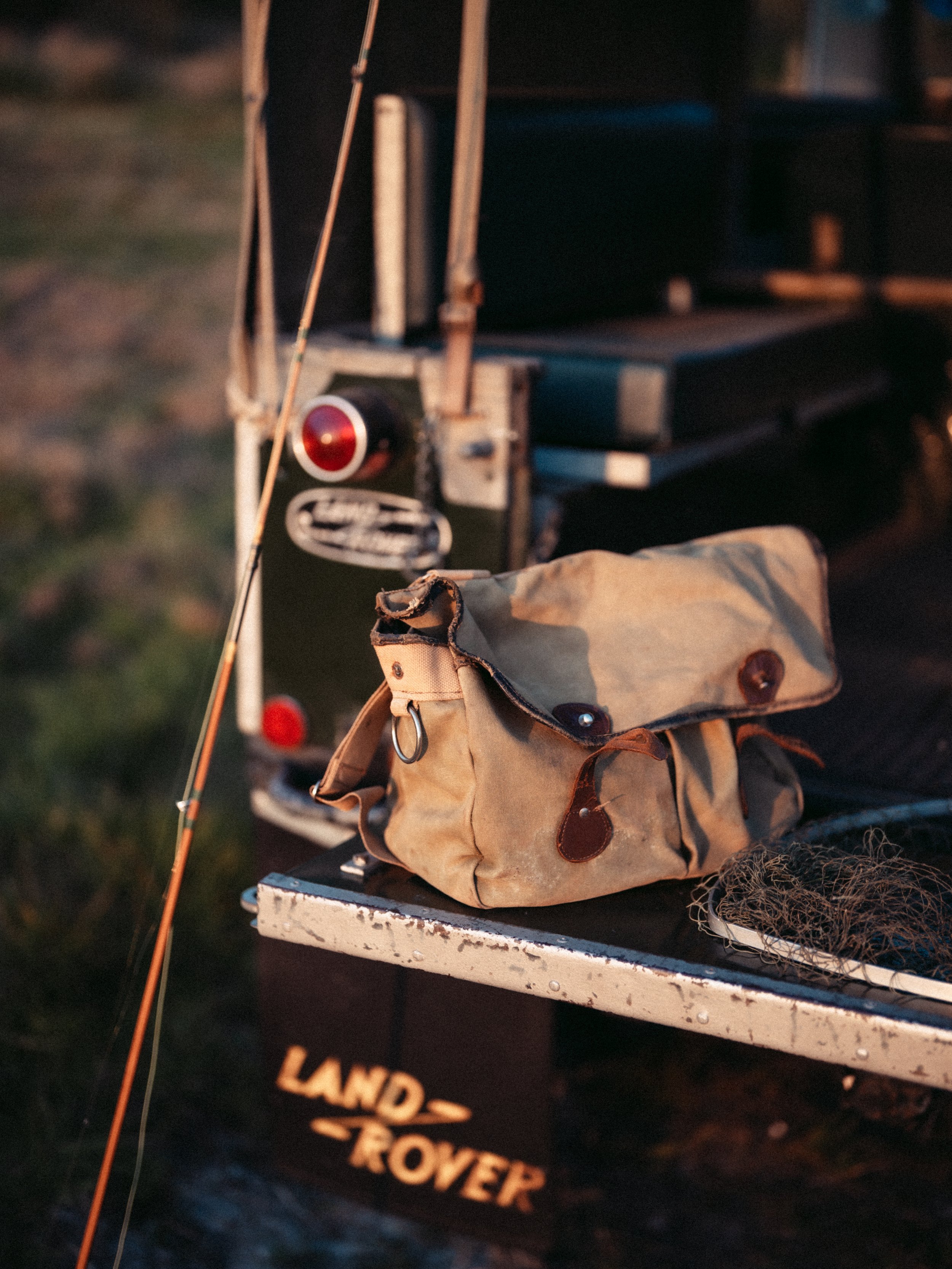Close-up of a camping or outdoor gear setup on a table, including a beige canvas bag, a fishing rod, a platform with wooden edges, and a dark background with the partially visible words 'LAND ROVER'.