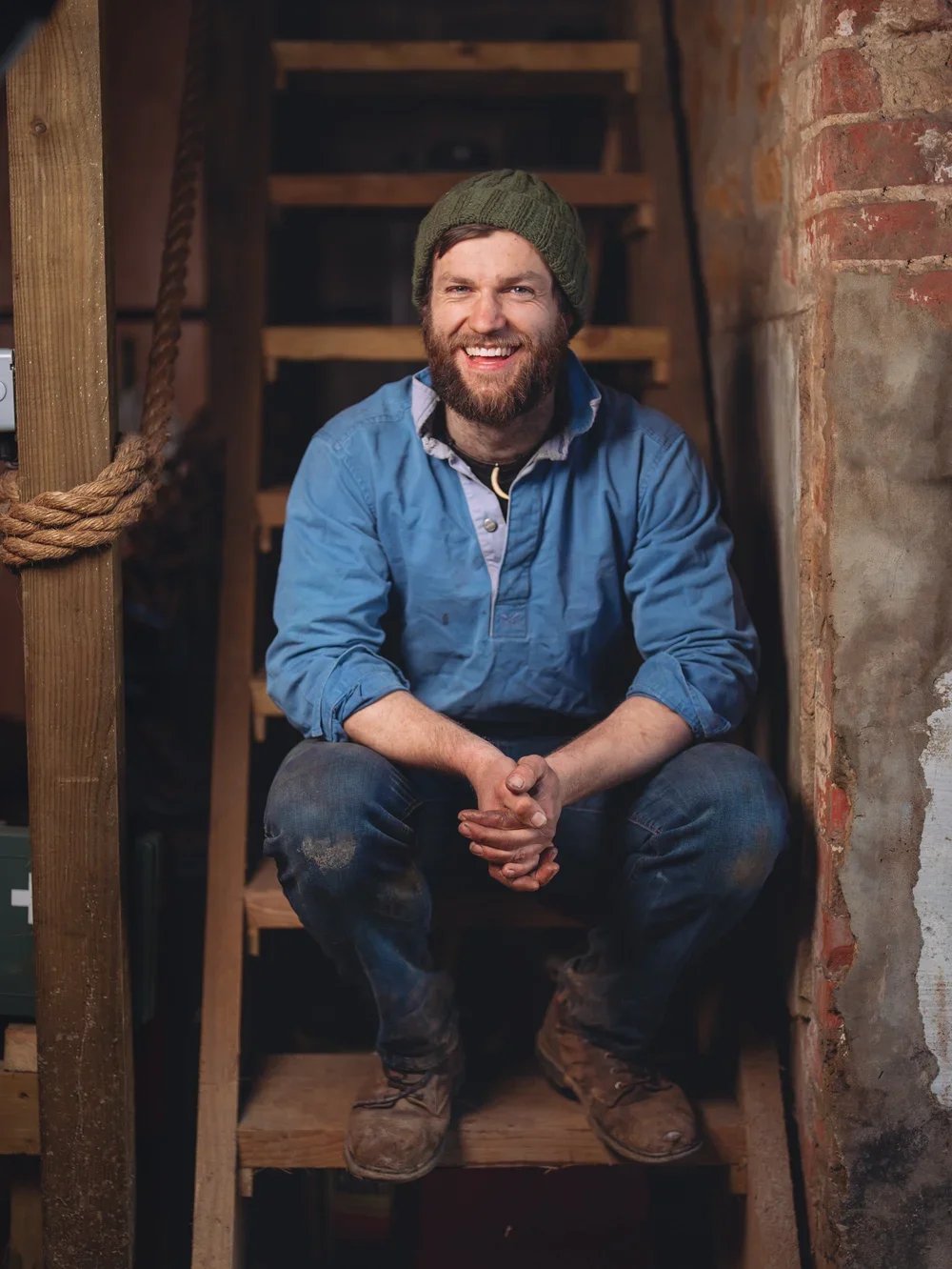 A man sitting on wooden stairs in a rustic setting, smiling, wearing a blue shirt, jeans, brown work boots, and a green beanie.