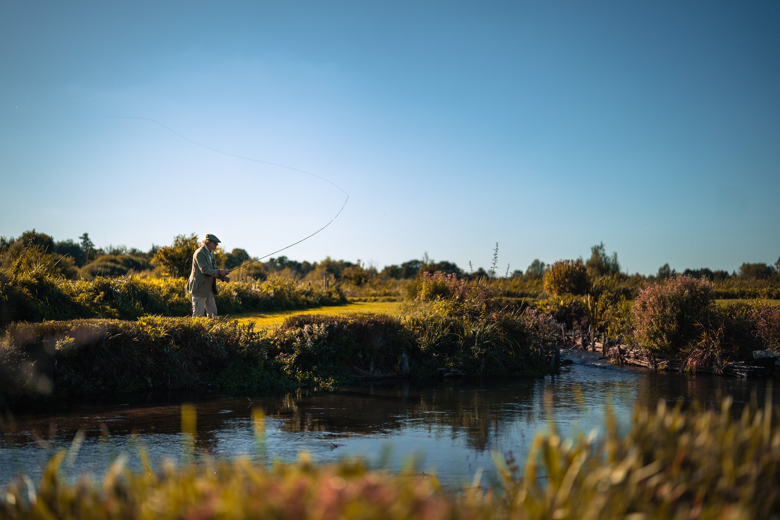 An older man fishing in a river near a grassy and bushy shoreline on a clear day.