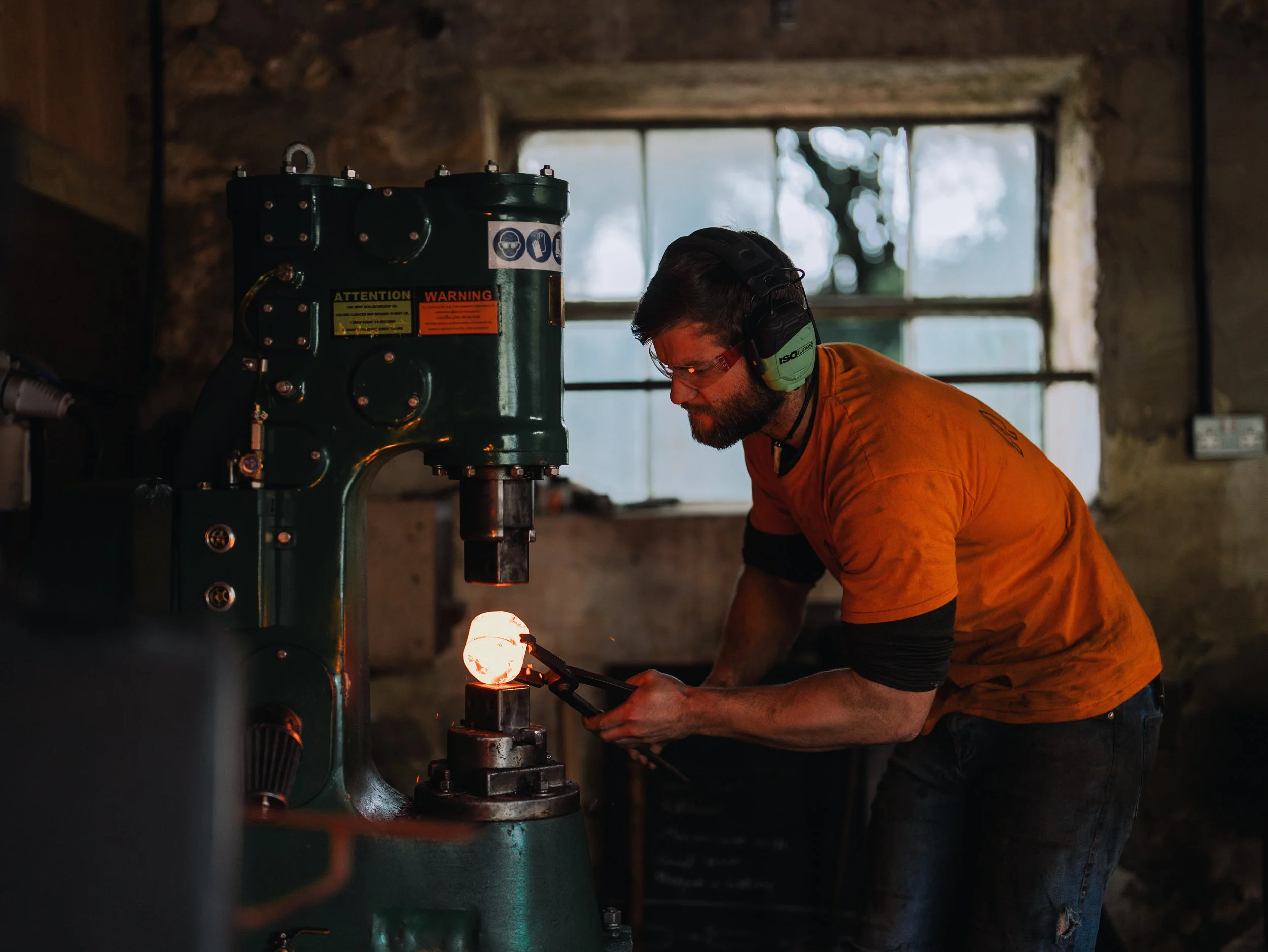 A man wearing safety glasses, ear protection, and an orange shirt leaning over a large vintage machine with a glowing metal object inside.