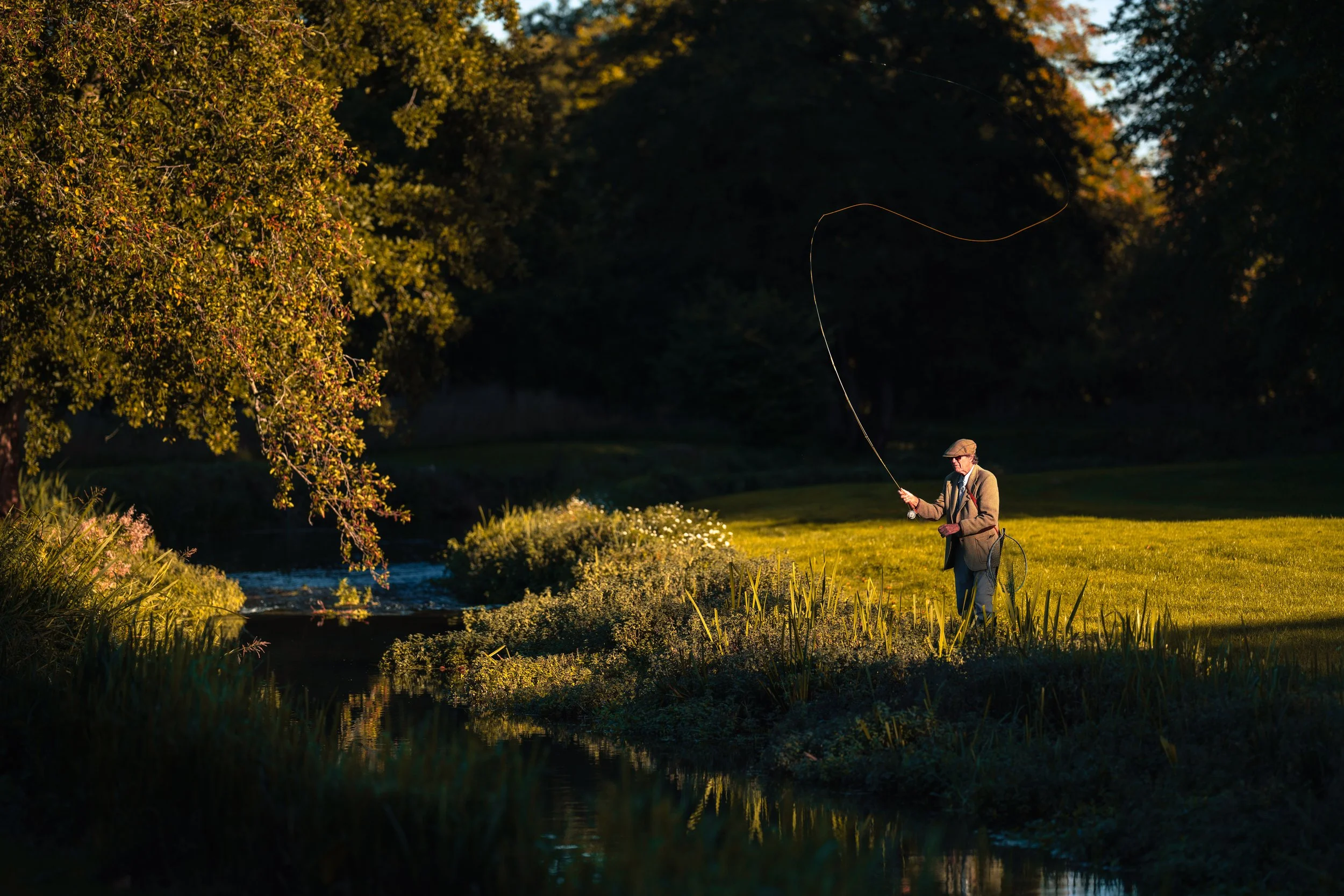 An elderly man fishing by a small river in a park during autumn, with trees showing fall colors in the background and the sun casting a golden light.