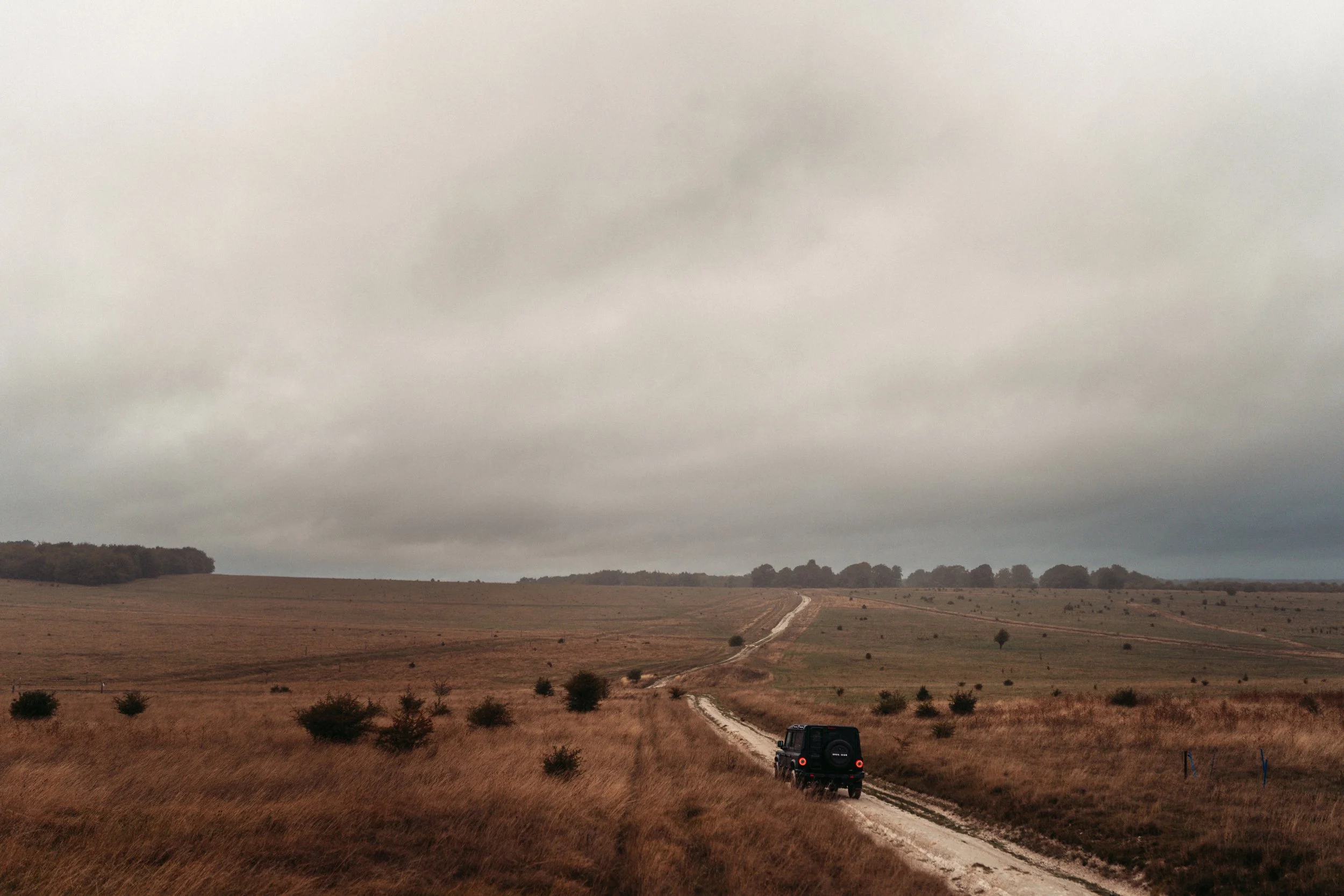 A black SUV driving on a winding dirt road through an open landscape with dry grass and a few scattered trees. The sky is overcast with gray clouds, and distant hills are visible on the horizon.