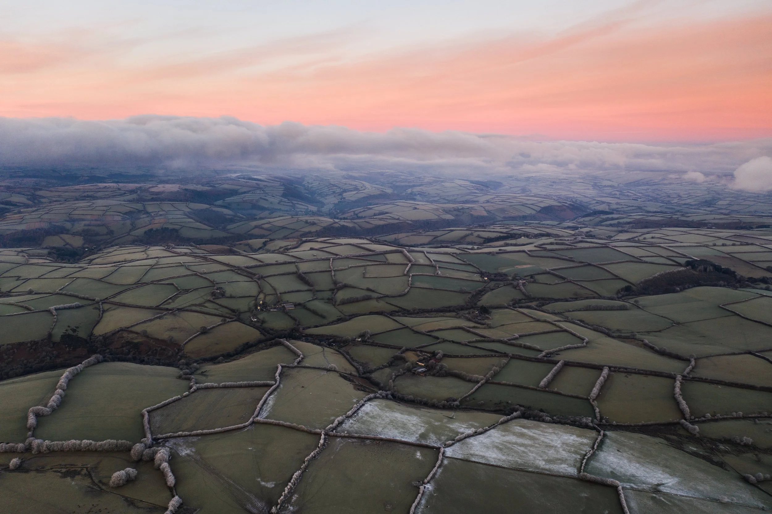 Aerial view of a landscape showing patchwork of fields divided by hedgerows or fences, with a cloudy sky and a pinkish sunset in the background.