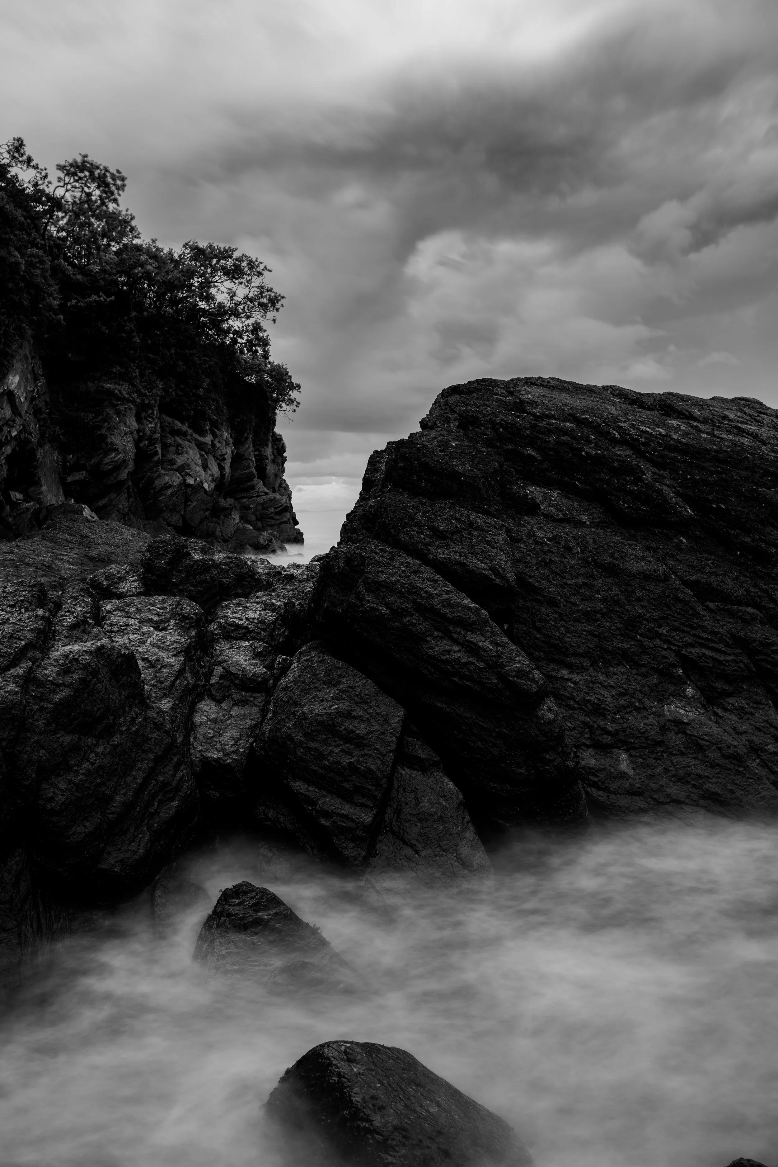 Black and white photo of rocky shoreline with large boulders and trees on the left, stormy clouds overhead, and water flowing around rocks.