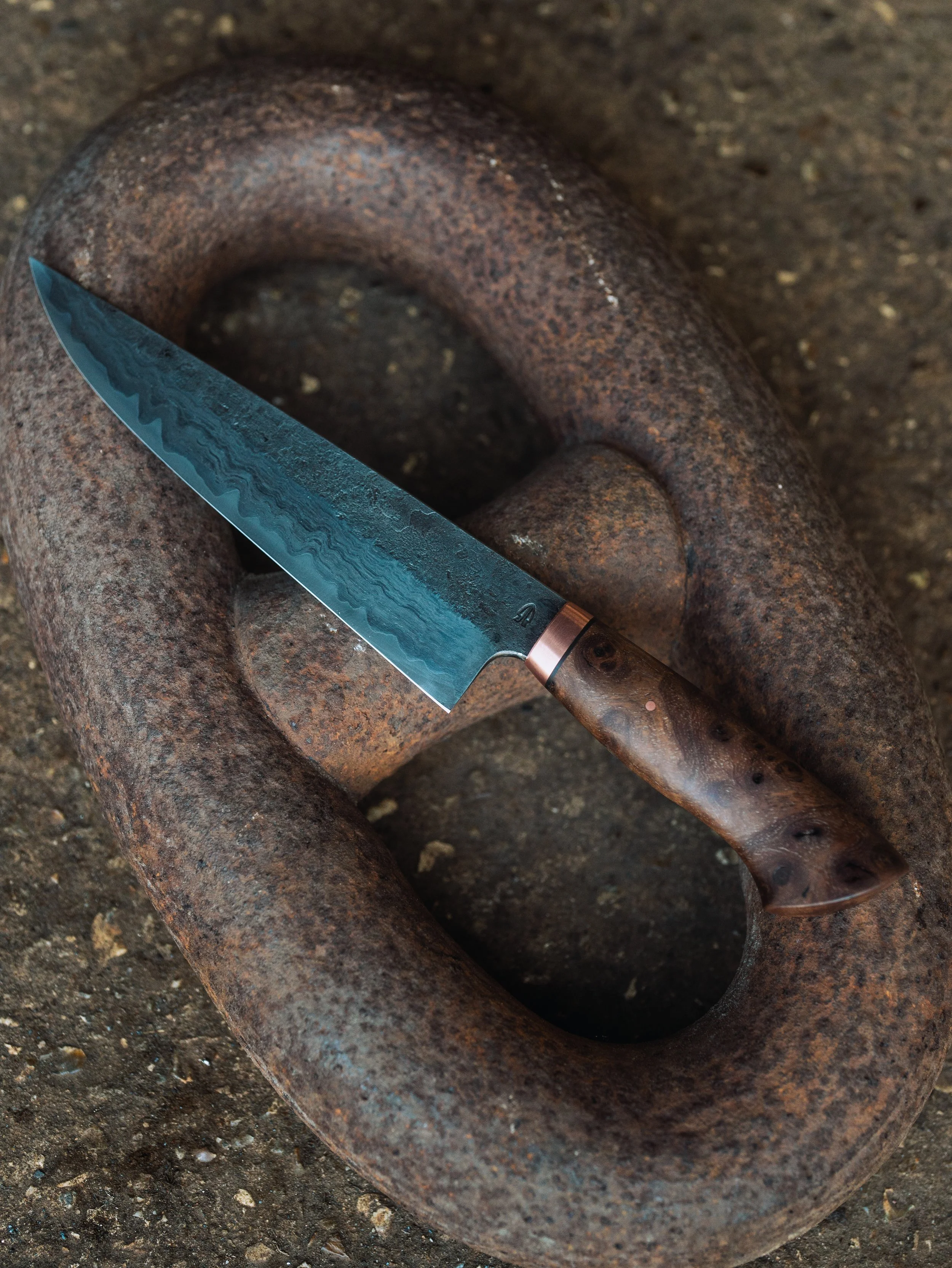 A rusty, circular metal object with a hole in the center, resting on a rough, dirt ground. A kitchen knife with a patterned blue blade and a brown handle is placed across the object.