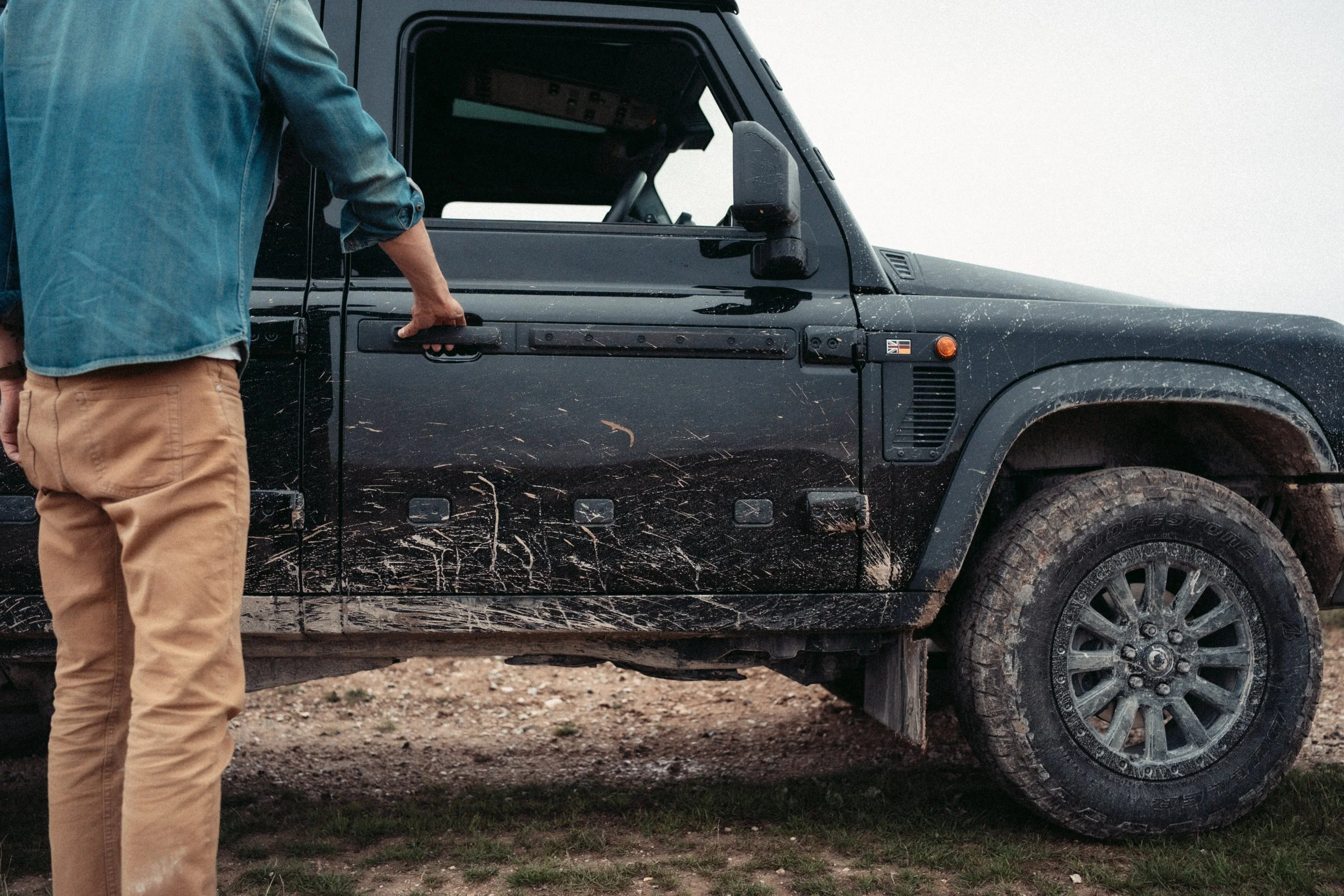 Person in denim jacket and tan pants holding a black handle on the door of a black and muddy off-road vehicle parked on a dirt and grass area.