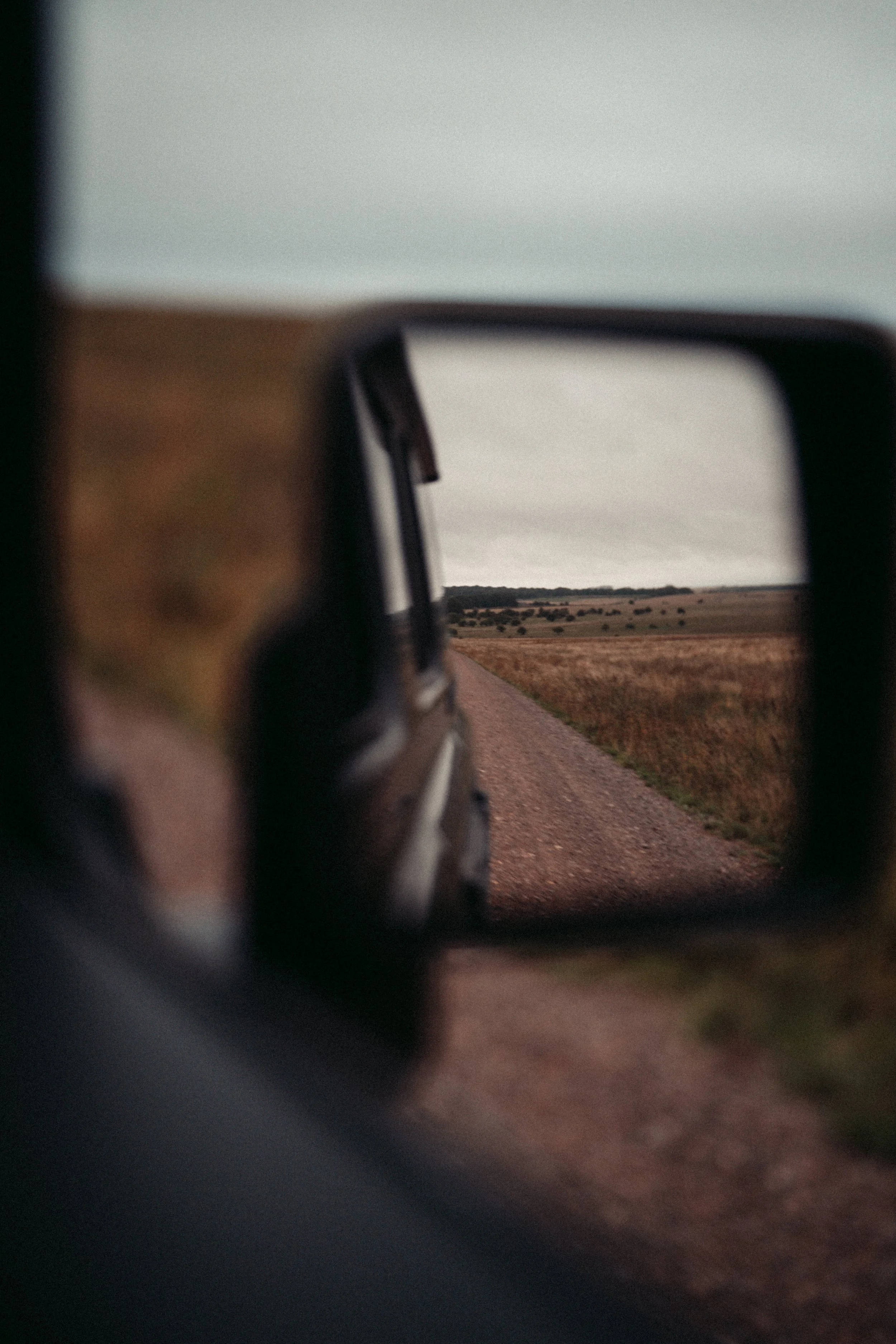 View of a rural landscape seen through a car's side mirror, showing an overcast sky and open fields.