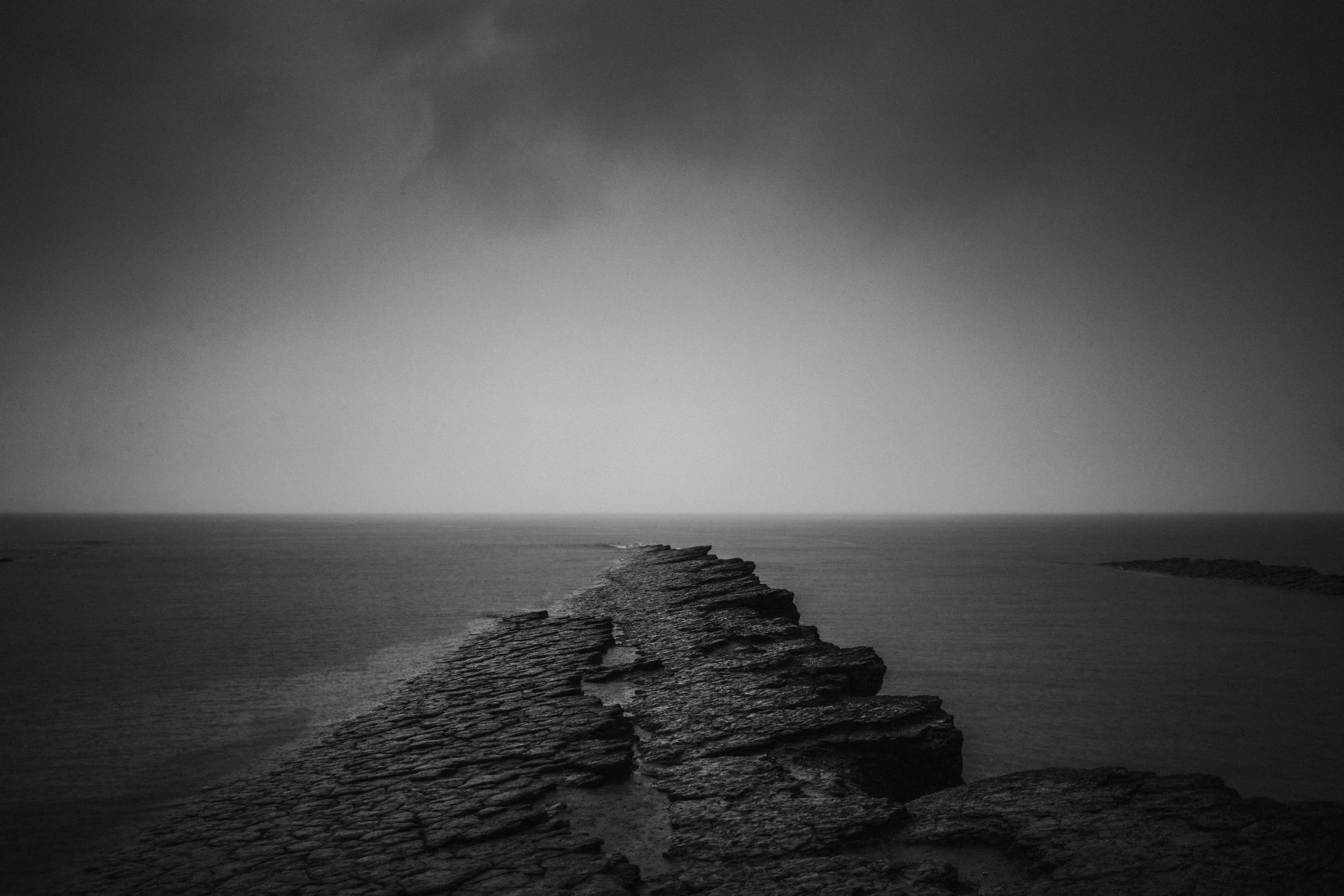 A black and white photograph of a rocky coastline with a narrow strip of beach extending into the ocean, under a dark sky with faint clouds.