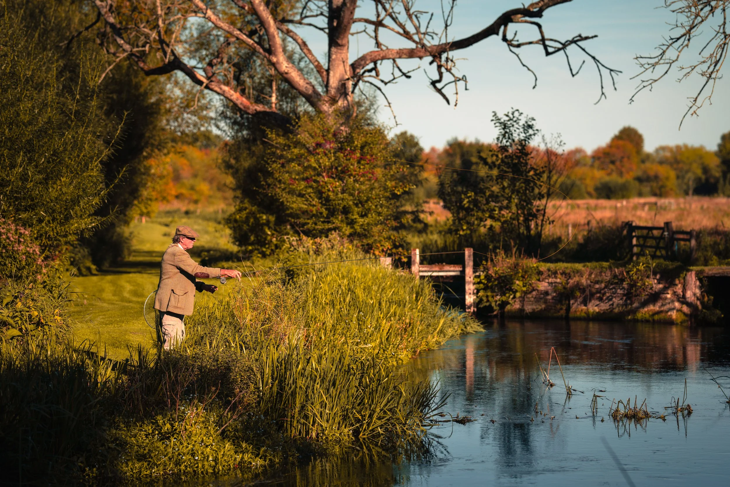 An elderly man in a tweed jacket and cap stands by a river, fishing with a fishing rod. The scene is set during the fall, with colorful trees and bushes along the riverbank and a clear blue sky overhead.