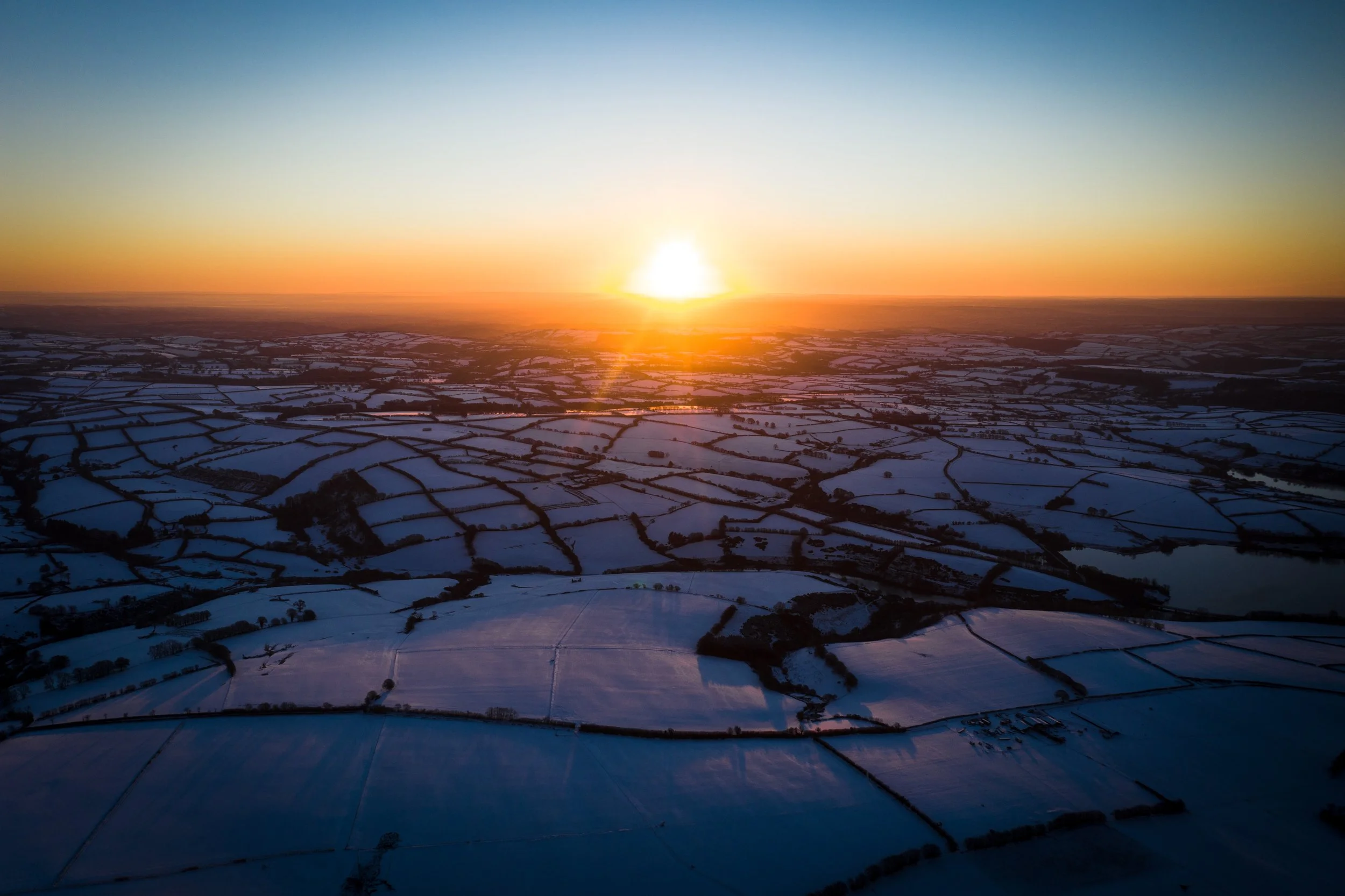 Aerial view of snow-covered fields and farmland at sunset with a clear sky.