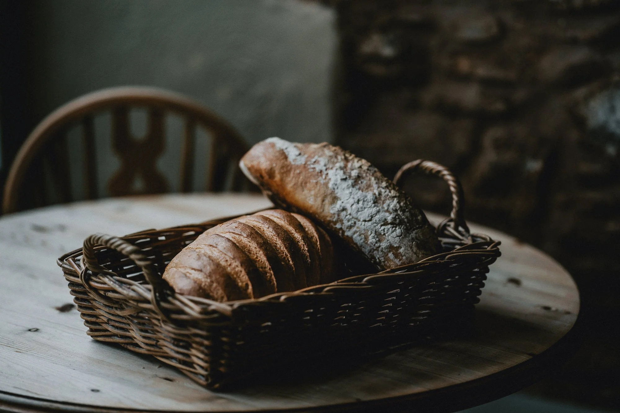 A basket of sliced bread and a loaf of bread dusted with flour on top, placed on a wooden surface in a rustic setting.