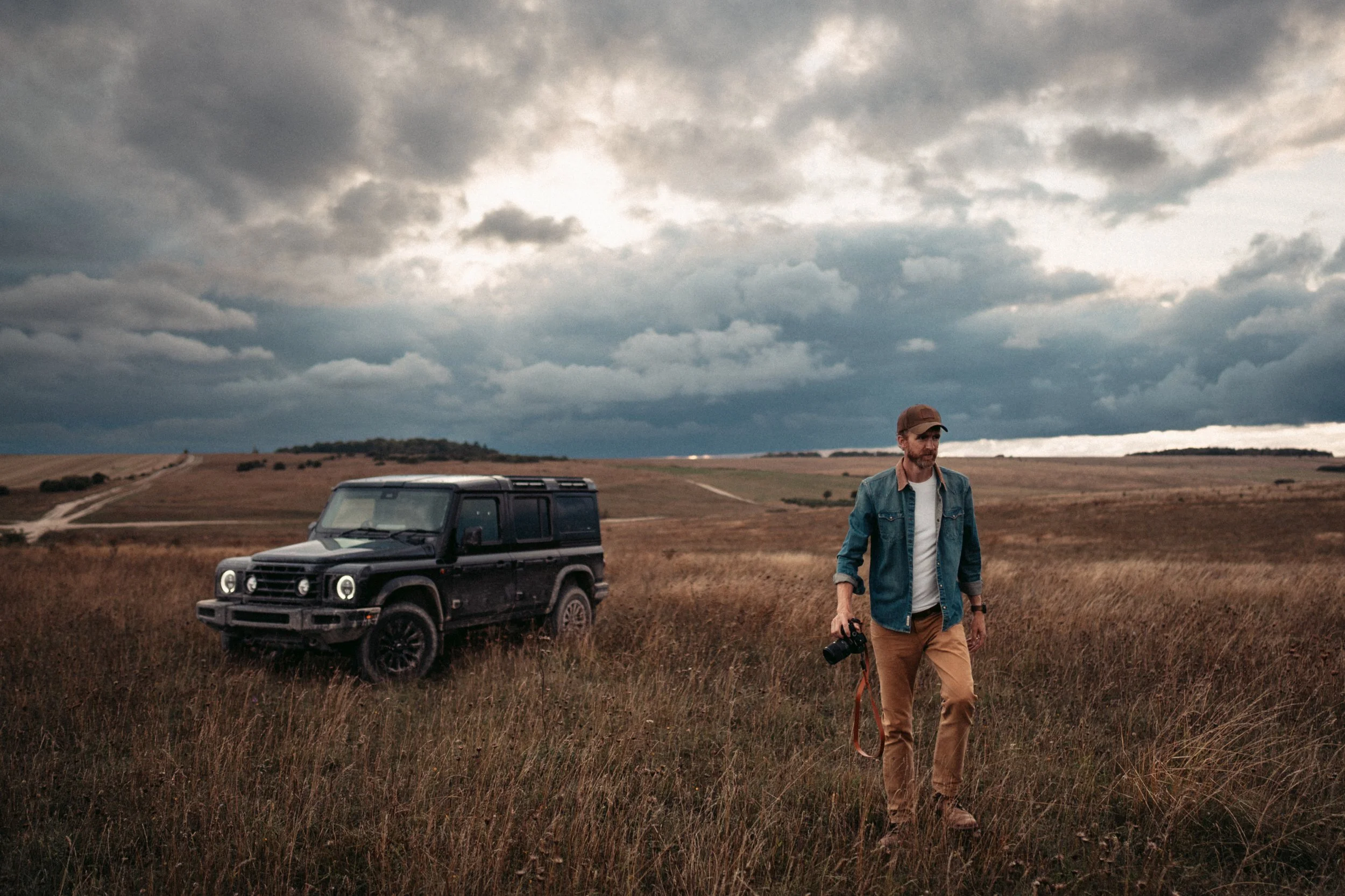 A man walking through a grassy field holding a camera, with a dark green SUV parked behind him and a cloudy sky overhead.
