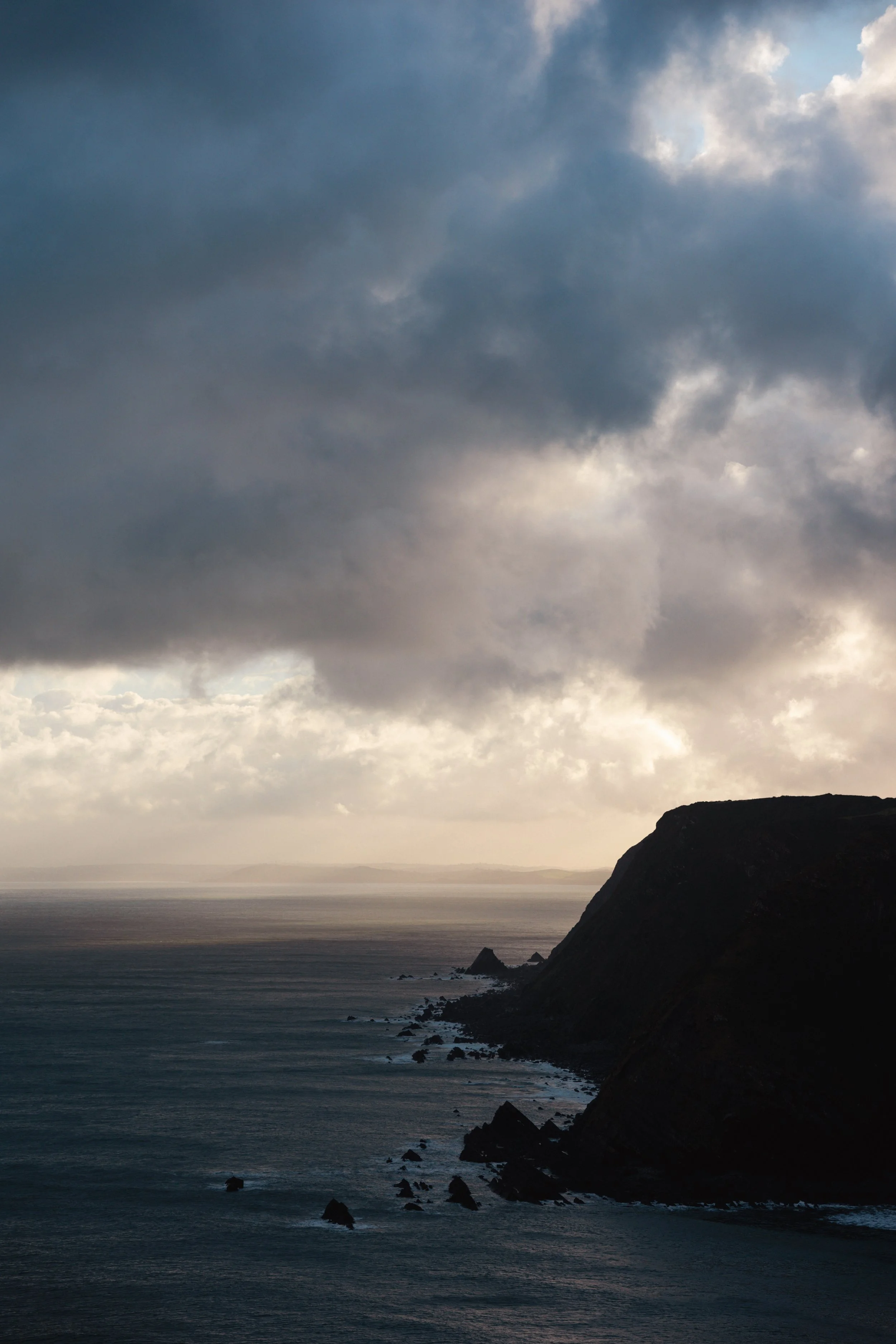 A coastal landscape featuring a dark, rocky headland on the right, extending into a calm sea. The sky is overcast with dark, dense clouds, with some light breaking through, creating a moody atmosphere.