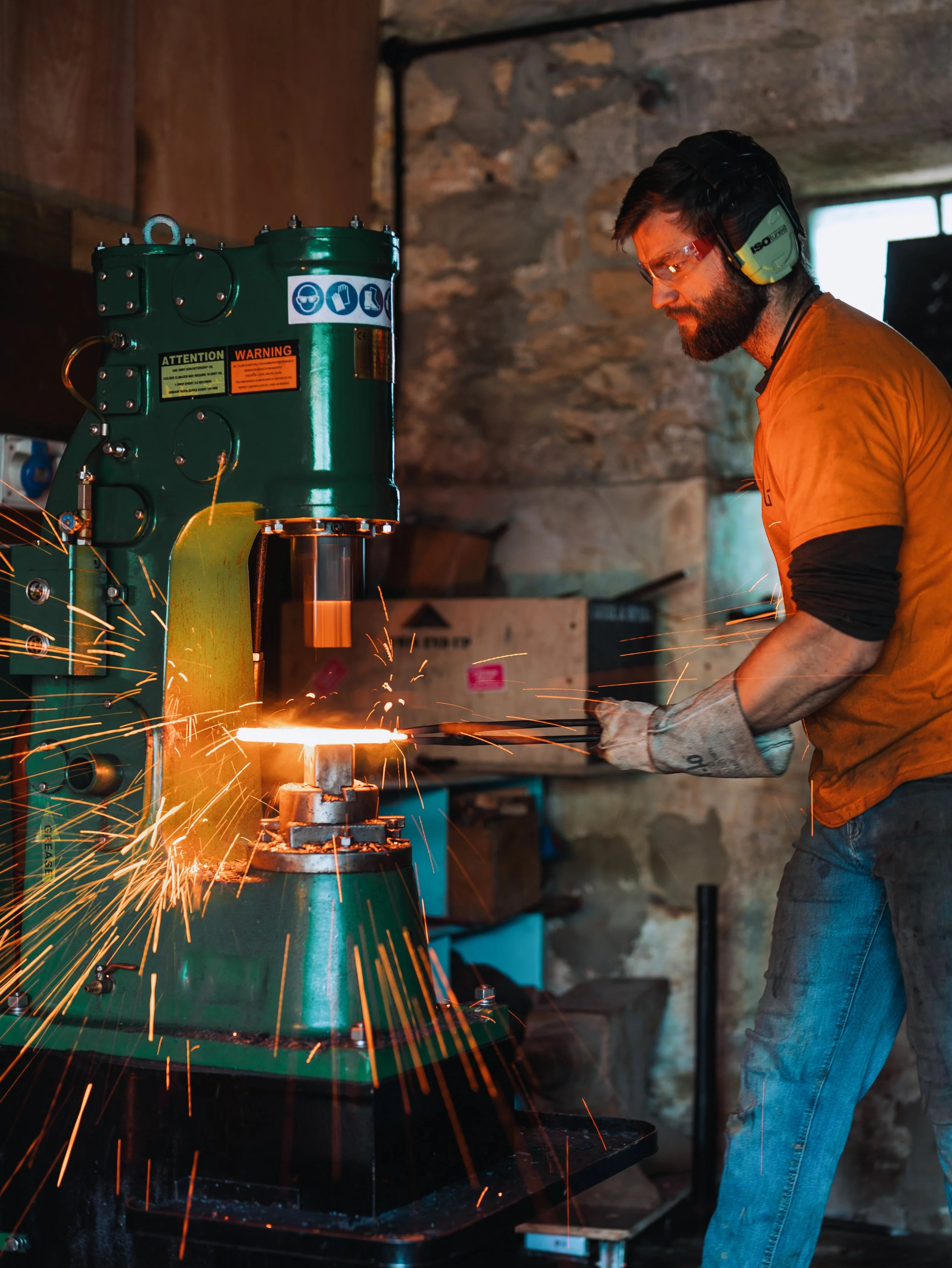 A man wearing safety goggles and ear protection is grinding a metal piece on a green industrial machine, producing sparks in a workshop with stone walls.