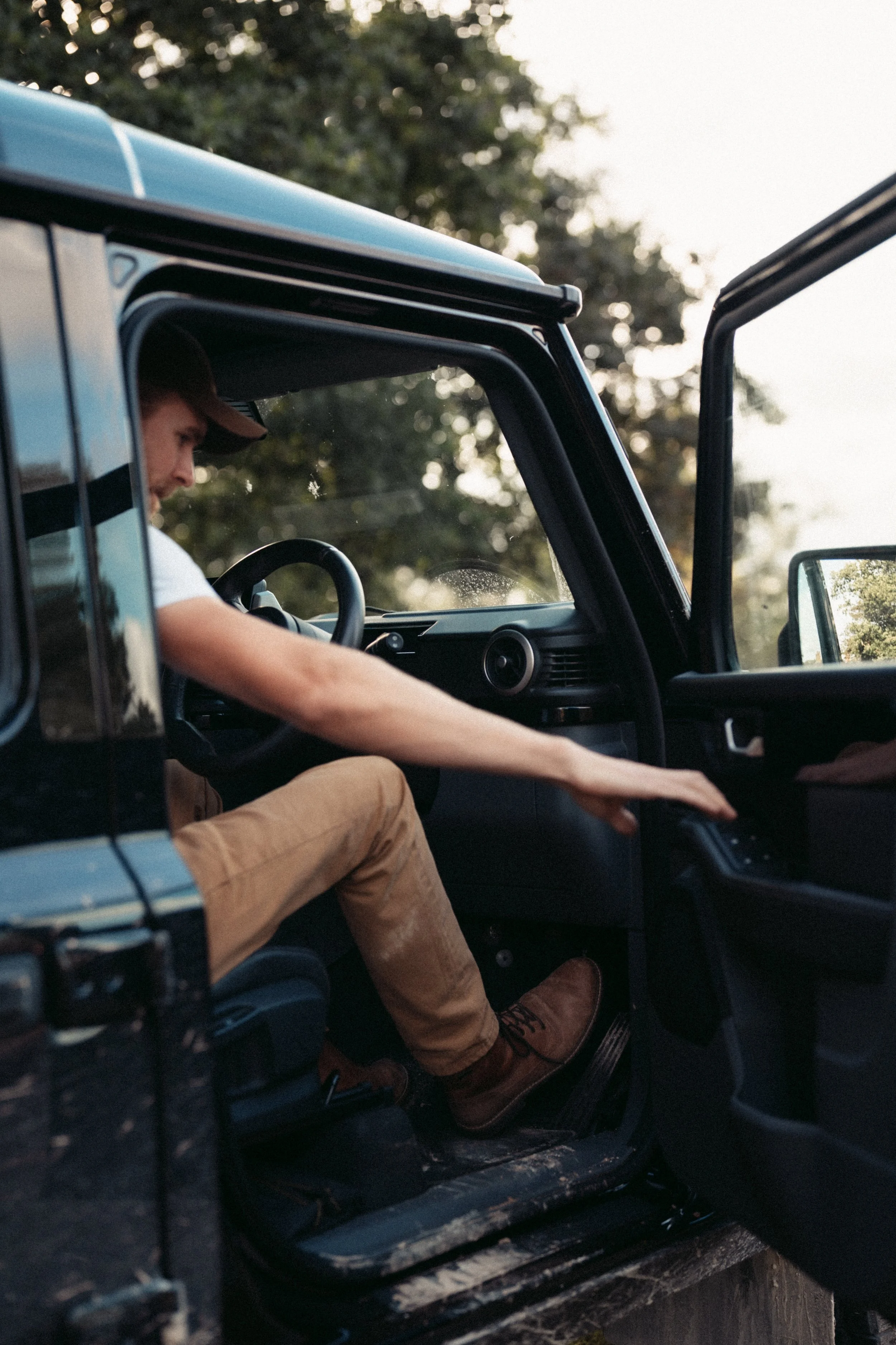 A person wearing a brown cap and tan boots is sitting inside a black vehicle, with the driver's side door open. The person is reaching toward the dashboard, and trees and sky are visible outside the vehicle.
