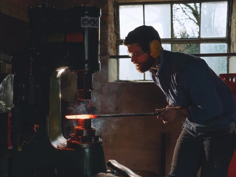 Man wearing yellow ear protection and glasses working with a forge, heating metal rod in a workshop with large windows in the background.