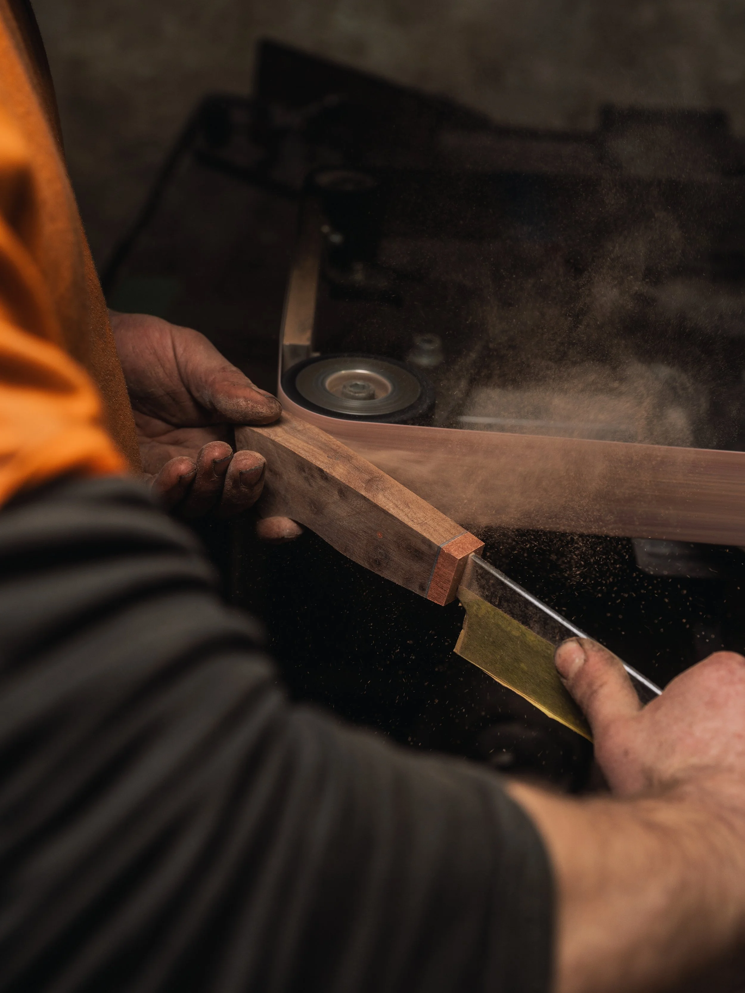 A person using a handheld power saw to cut a piece of wood in a woodworking workshop. The person's hands are visible, and the saw is actively cutting through the wood, producing sawdust.