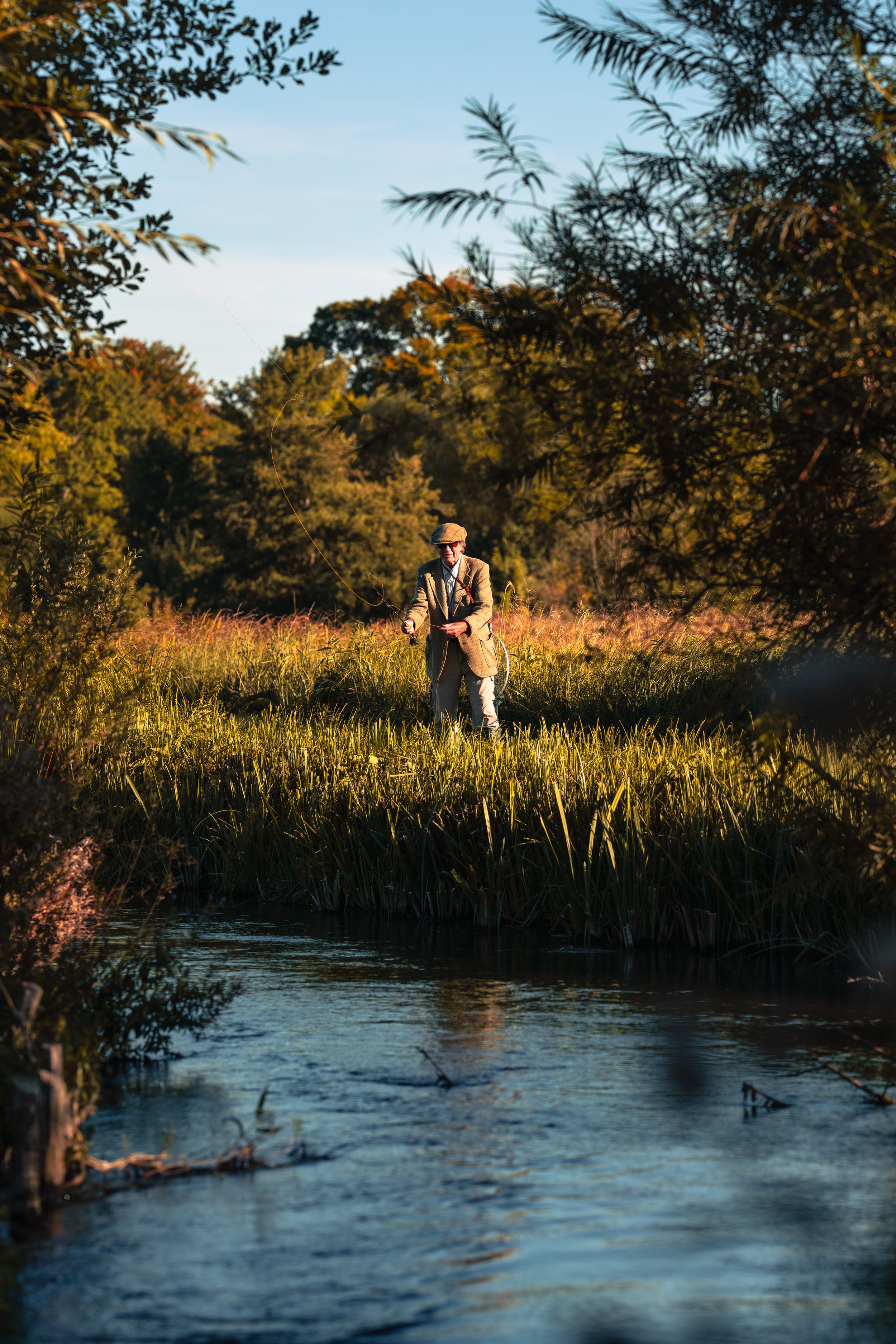 An older man with a hat and glasses standing in a grassy area by a stream, holding a fishing rod with a line cast into the water, surrounded by trees and foliage on a sunny day.