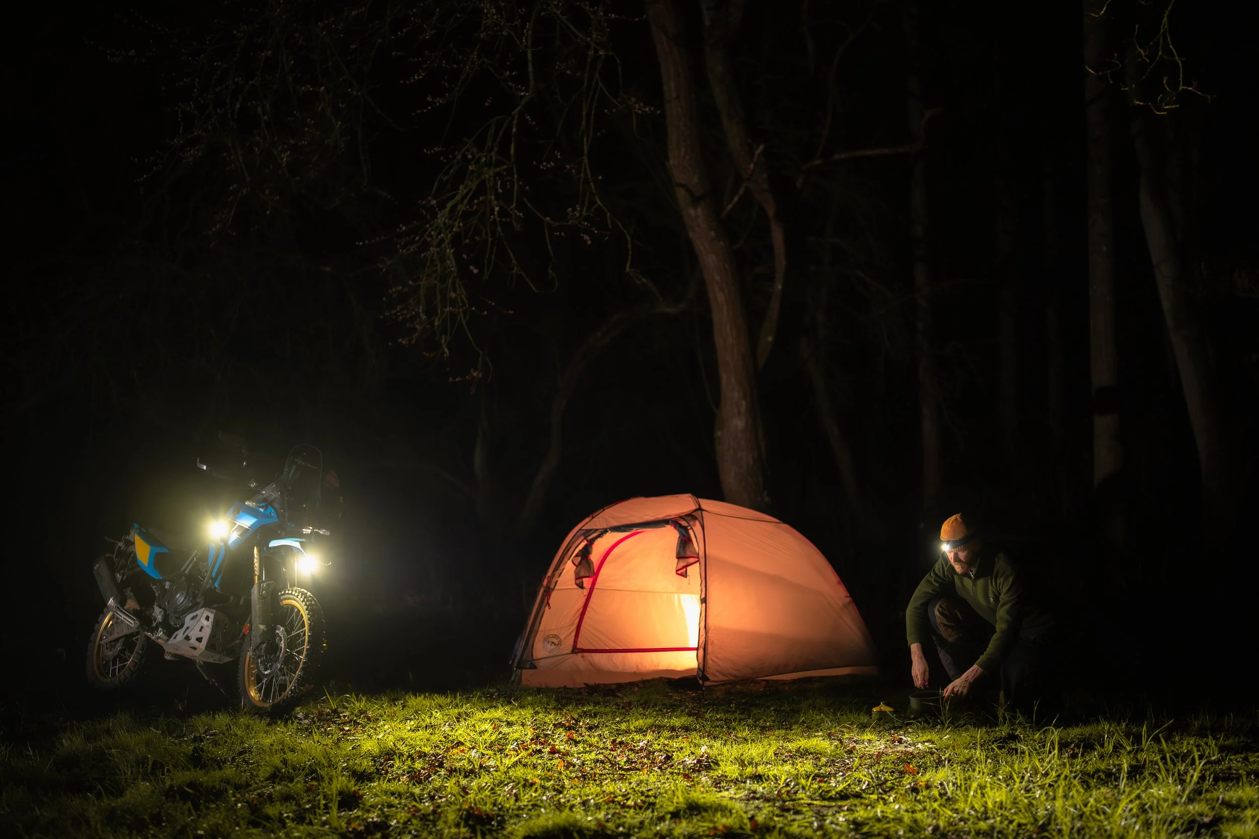 Night camping scene with a person setting up a tent, a motorcycle with lights on, and trees in the background.