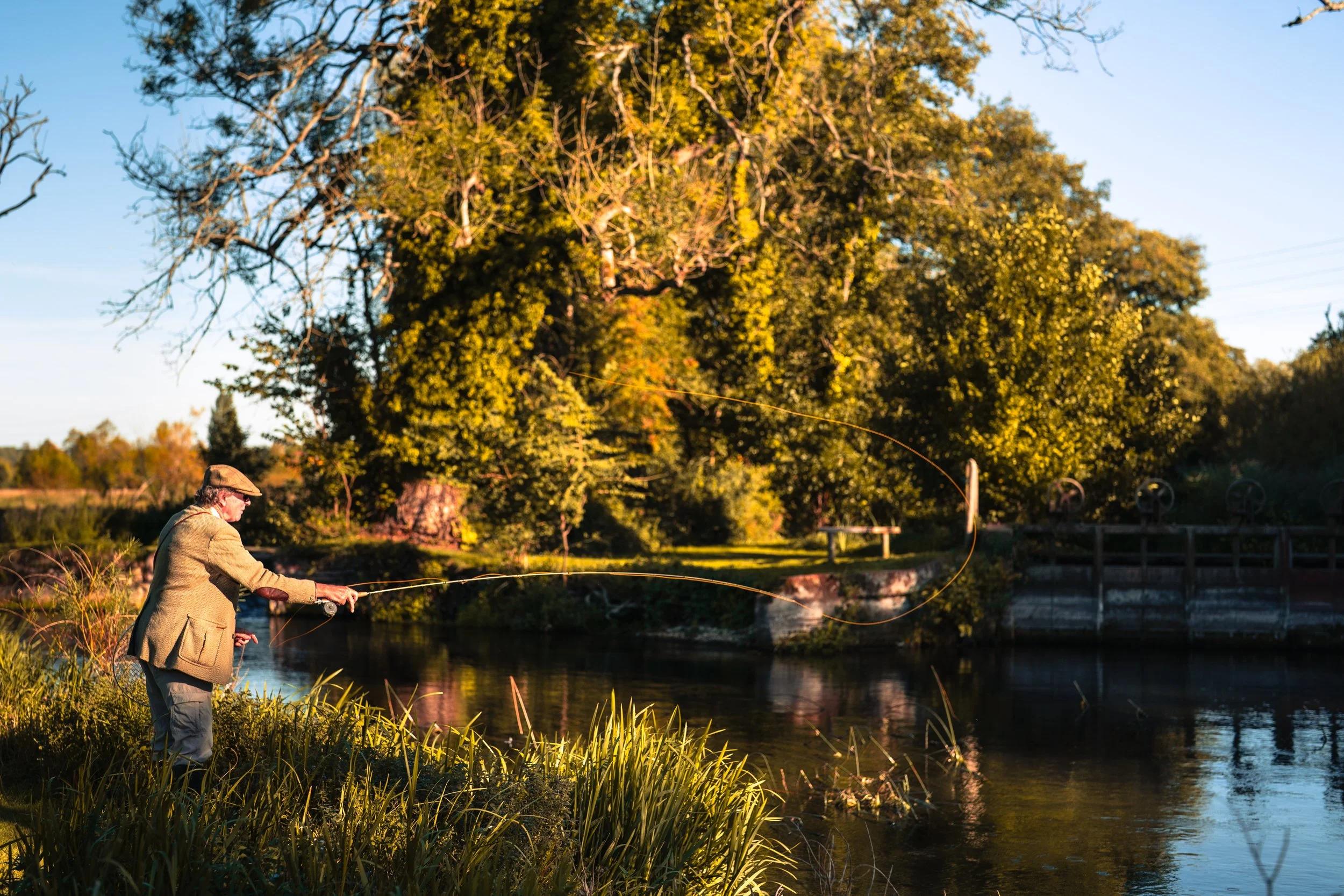 An elderly man wearing a jacket and hat fishing by a river at sunset, with large green trees in the background.