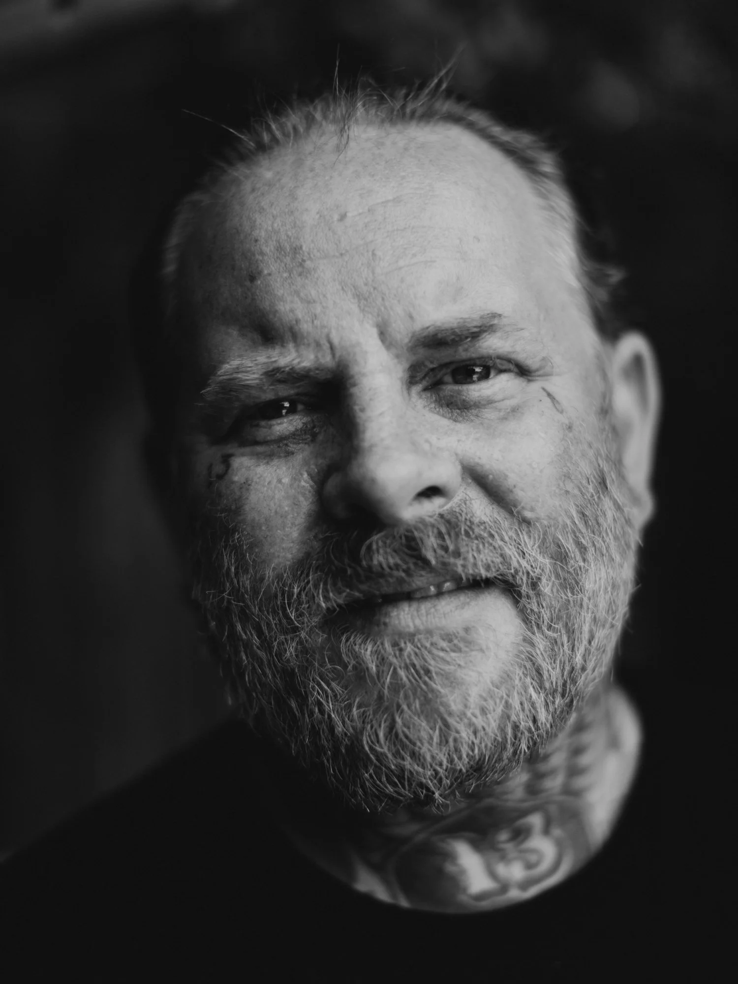Close-up black-and-white photograph of a person's face, showing the nose, eyes, and forehead, with a partially visible tattoo on the neck.