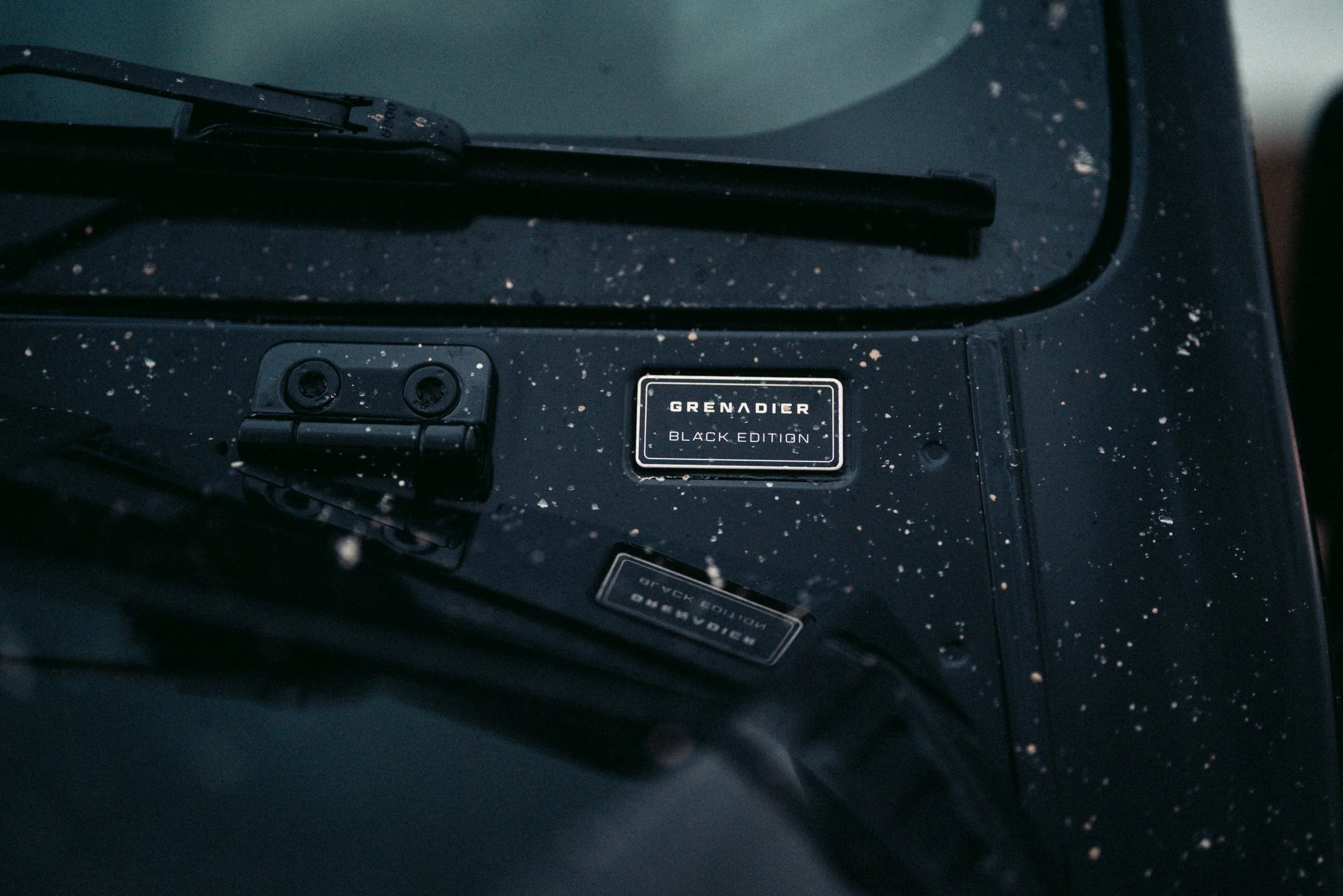 Close-up of a black device with a label that reads 'GRENADIER BLACK EDITION' and a black panel with screws, with small specks of dirt or dust on the surface.