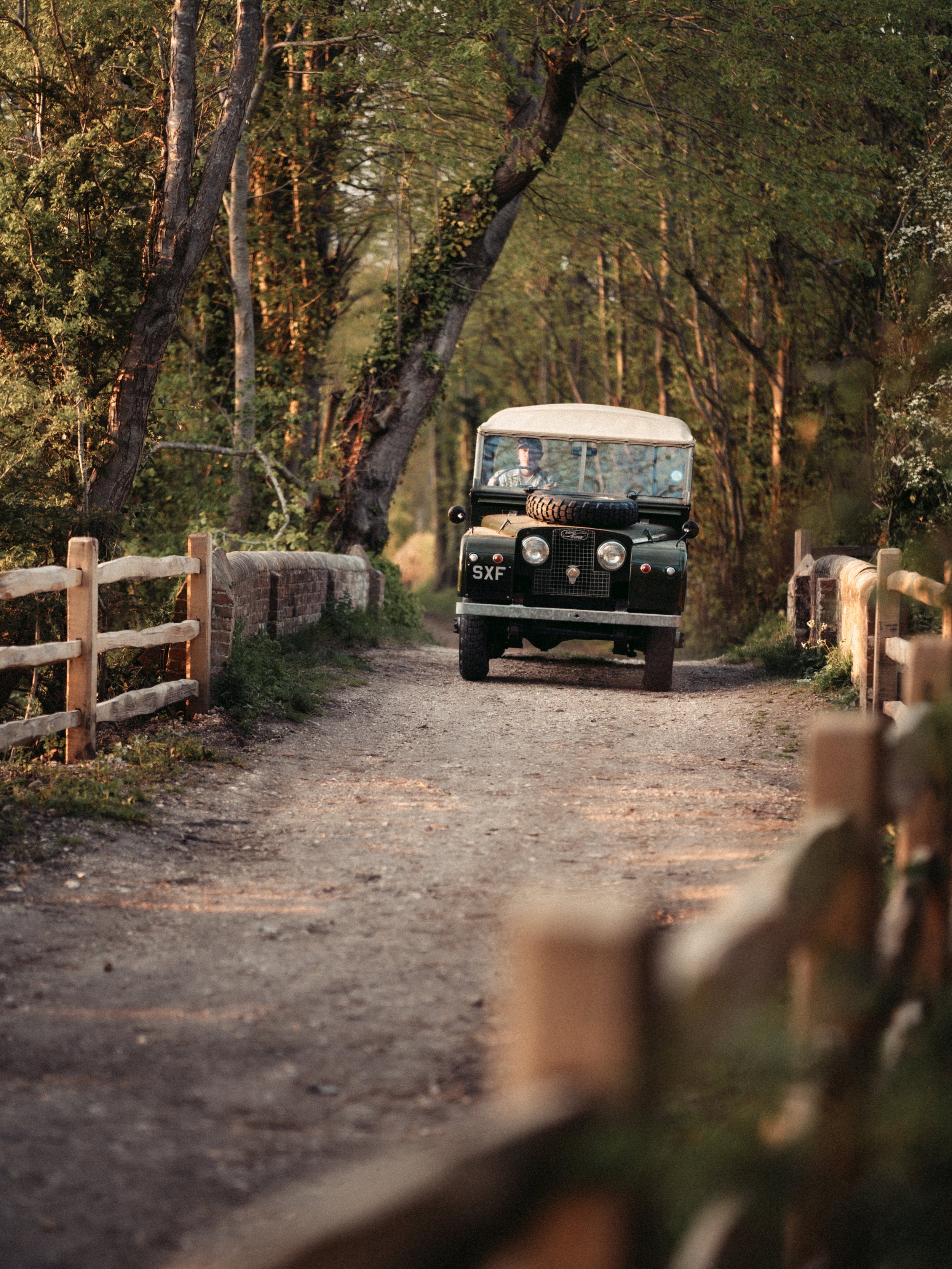 A vintage black Land Rover with a spare tire on the front, driving down a dirt road surrounded by trees during sunset.