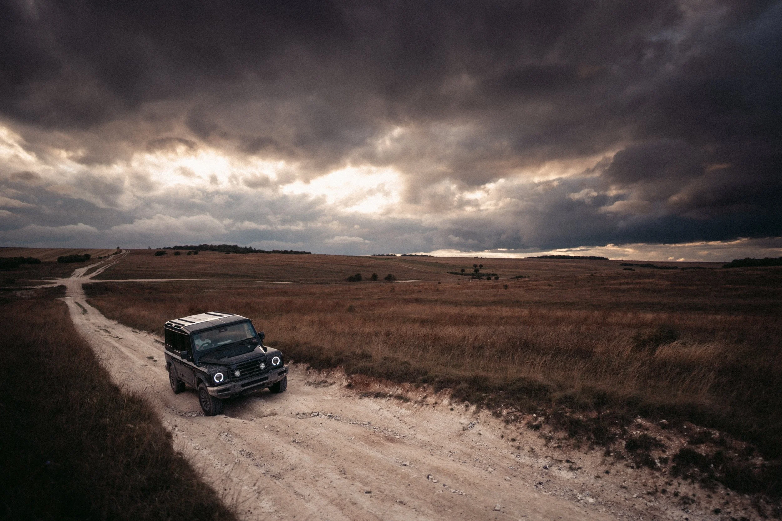 A black off-road vehicle driving on a dirt road through a grassy landscape under a dark stormy sky.