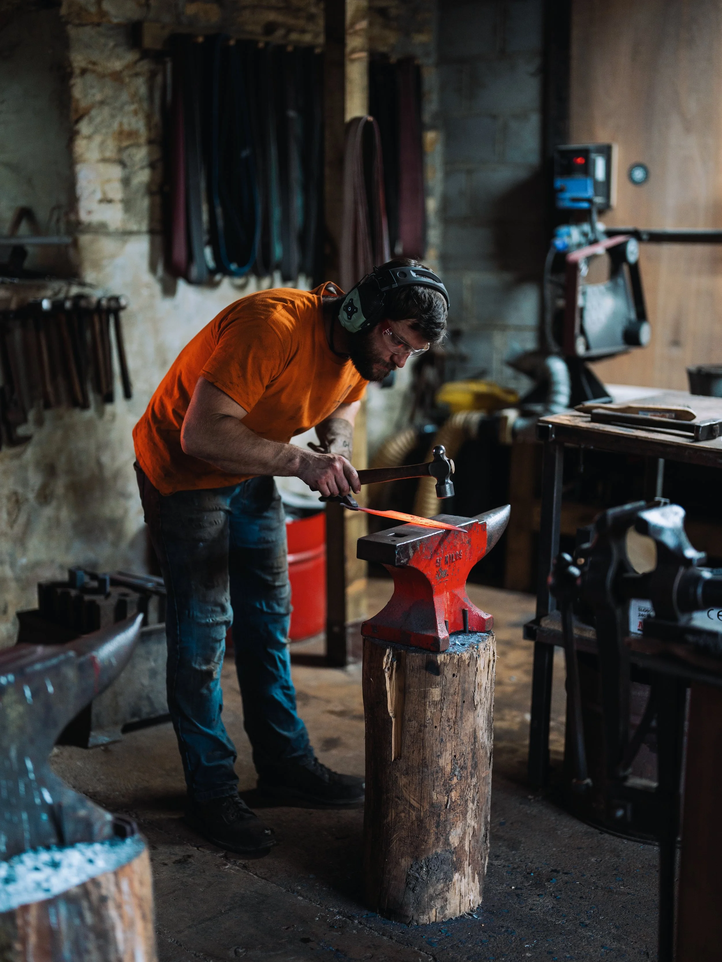 A man in an orange shirt and safety glasses, wearing a black helmet, is hammering a piece of metal on an anvil in a woodworking or blacksmithing workshop. The background shows hooks with hanging tools and equipment, and a workbench with various tools