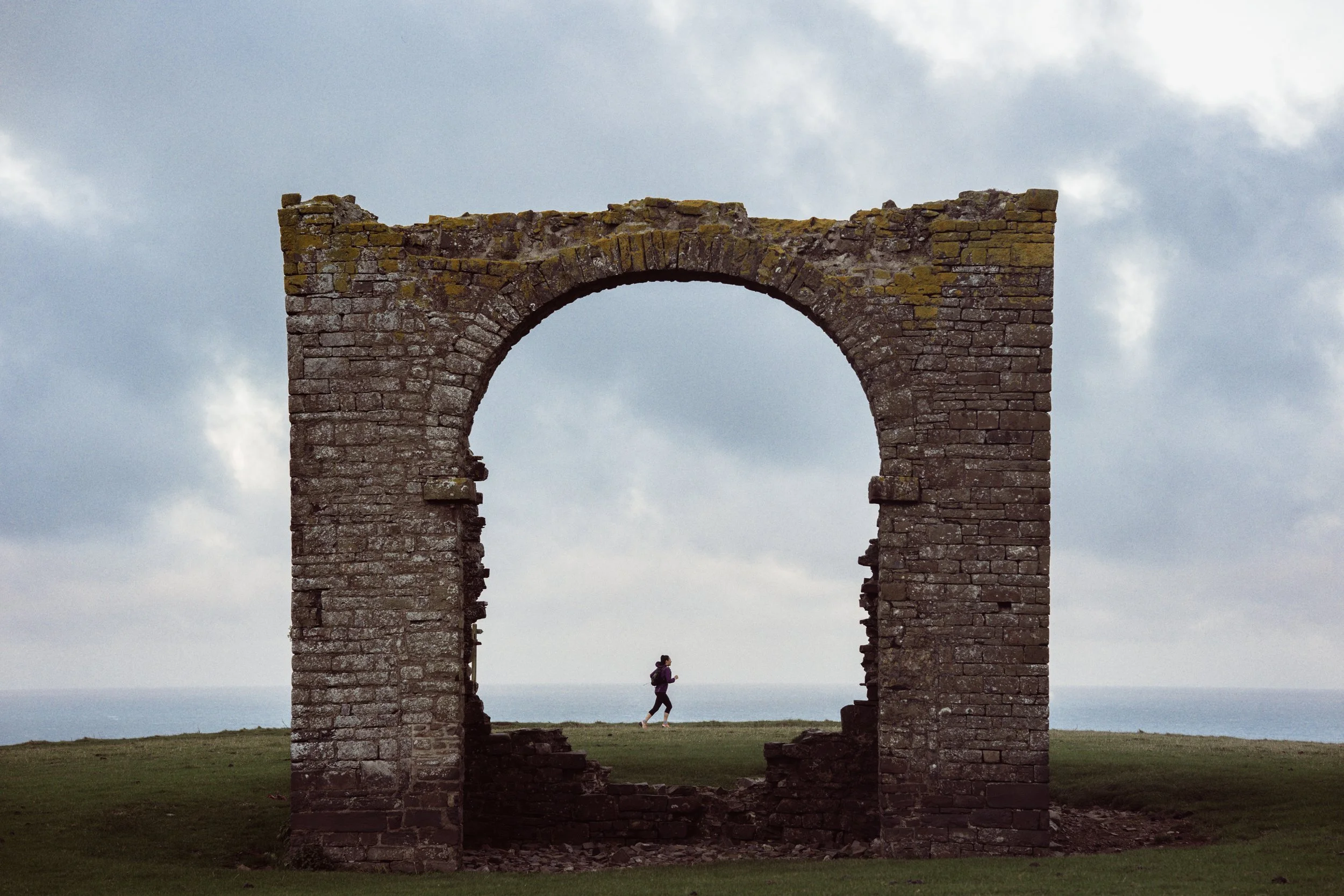 A person walking past ancient stone ruins with a large arch, overlooking a cloudy sky and distant landscape.