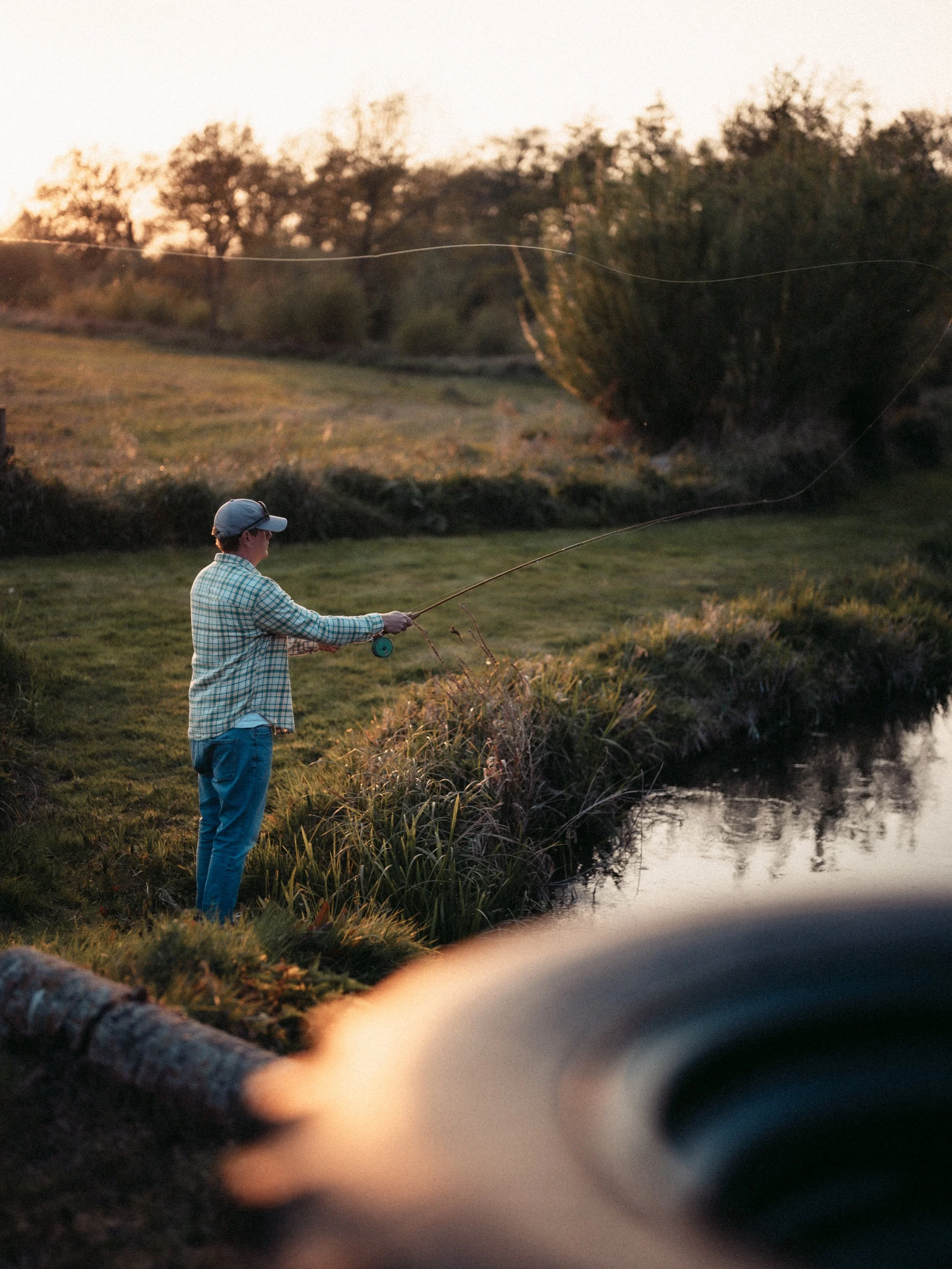 A man standing by a riverbank fishing at sunset, wearing a plaid shirt and a baseball cap.