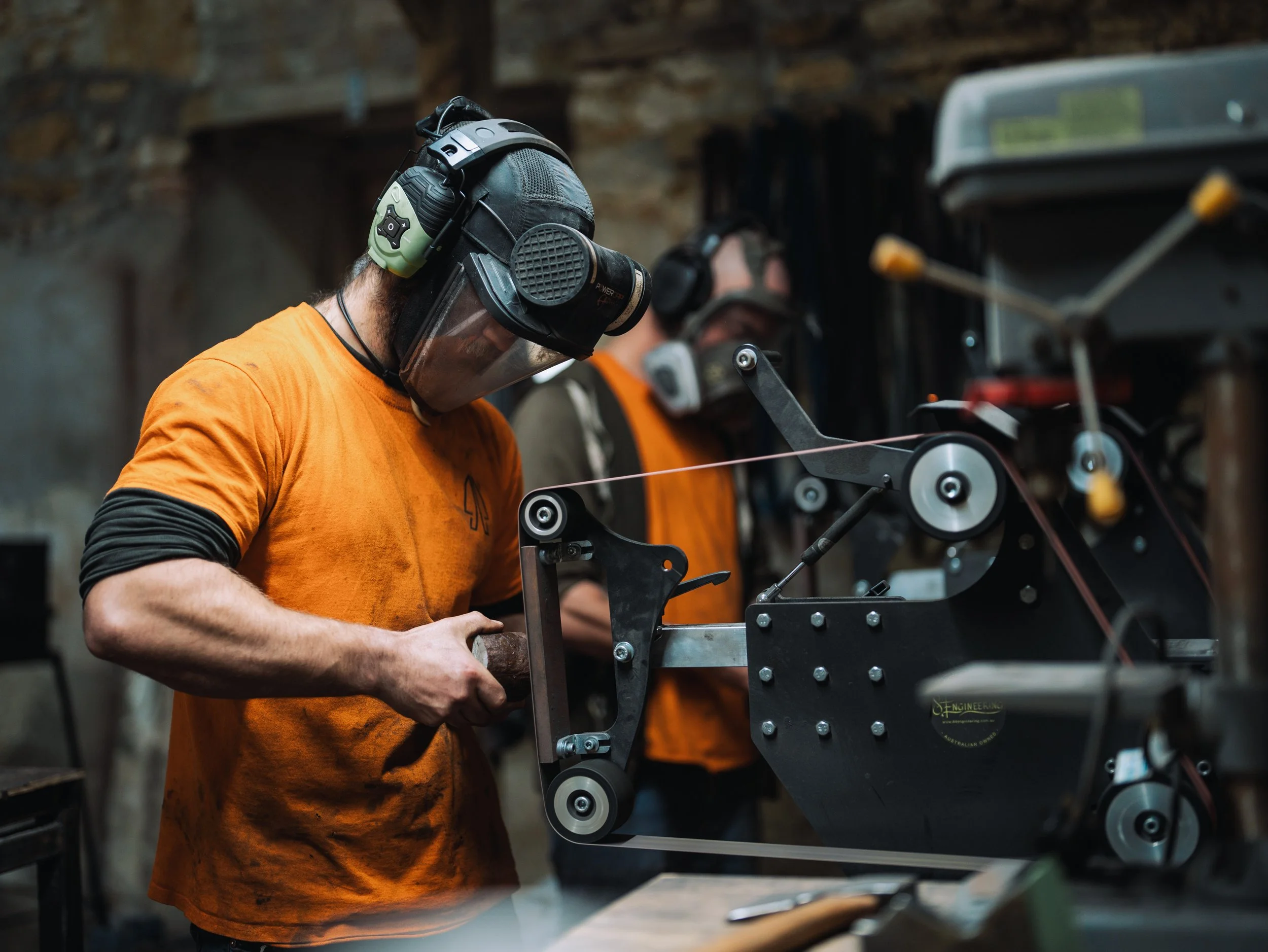 A person in an orange shirt working on a mechanical or engineering project in a workshop, with another person in the background also working on equipment.