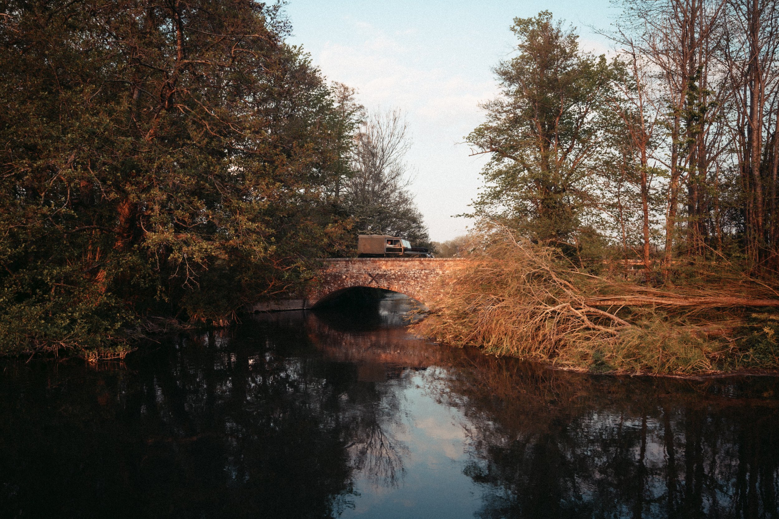 A small stone bridge crossing a calm river, surrounded by dense trees with autumn foliage, and a vintage truck parked on the bridge.